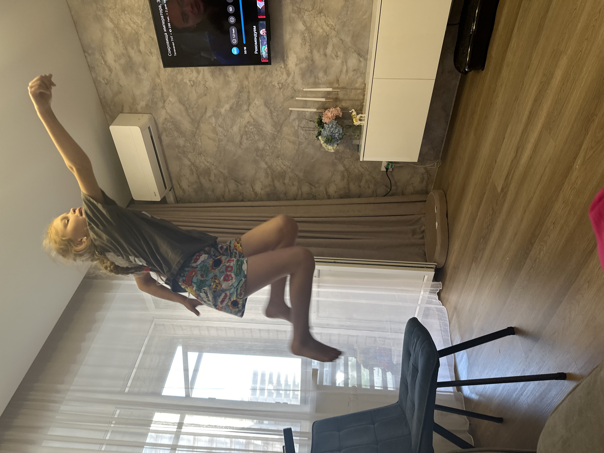 This media file shows a young girl mid-air, appearing to be jumping or flying in a living room. She is wearing a grey t-shirt and colorful patterned shorts. Her arms are outstretched, and her legs are bent as if she's in the middle of a dynamic movement. Her blonde hair is braided and hangs down her back.
The setting is a well-lit room with neutral-colored walls and wooden flooring. A large television screen is mounted on the wall, displaying text related to "Сокровища императора" and "Телеканал ТНТ" (Treasure of the Emperor, TNT Channel). There is a white cabinet below the TV with some decorative items, including candles and a small bouquet of flowers. An air conditioner is mounted on the wall above the cabinet. Sheer curtains are visible in the background, suggesting it might be daytime. A dark grey upholstered chair is positioned in the foreground to the right, with its legs extending towards the wooden floor.
The overall impression is one of playfulness and exuberance. The child's pose and the frozen motion suggest a moment of fun and energetic activity within a domestic environment. The visible text on the TV screen provides context about the media being watched, possibly influencing the child's imaginative play.