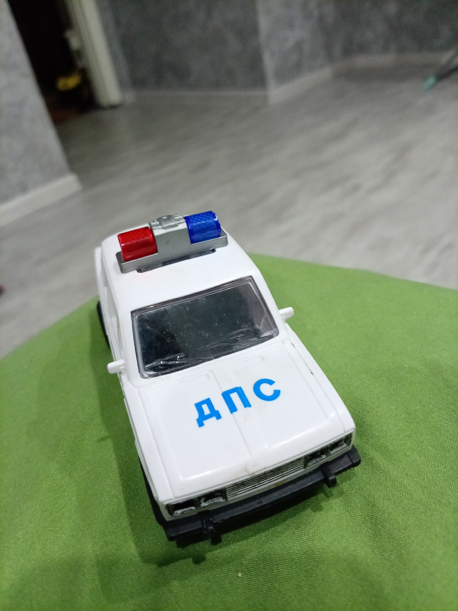 A white toy police car with blue lettering and a red and blue flashing light bar is positioned on a green fabric surface. The car is facing slightly to the right, with its hood angled towards the viewer. The background consists of a blurred indoor environment with gray walls and a light-colored floor, possibly a hallway or room. The lighting is dim and appears to come from overhead. The image is a close-up shot, focusing on the toy car. There is no indication of the specific location within Oktyabrsk, Russia.