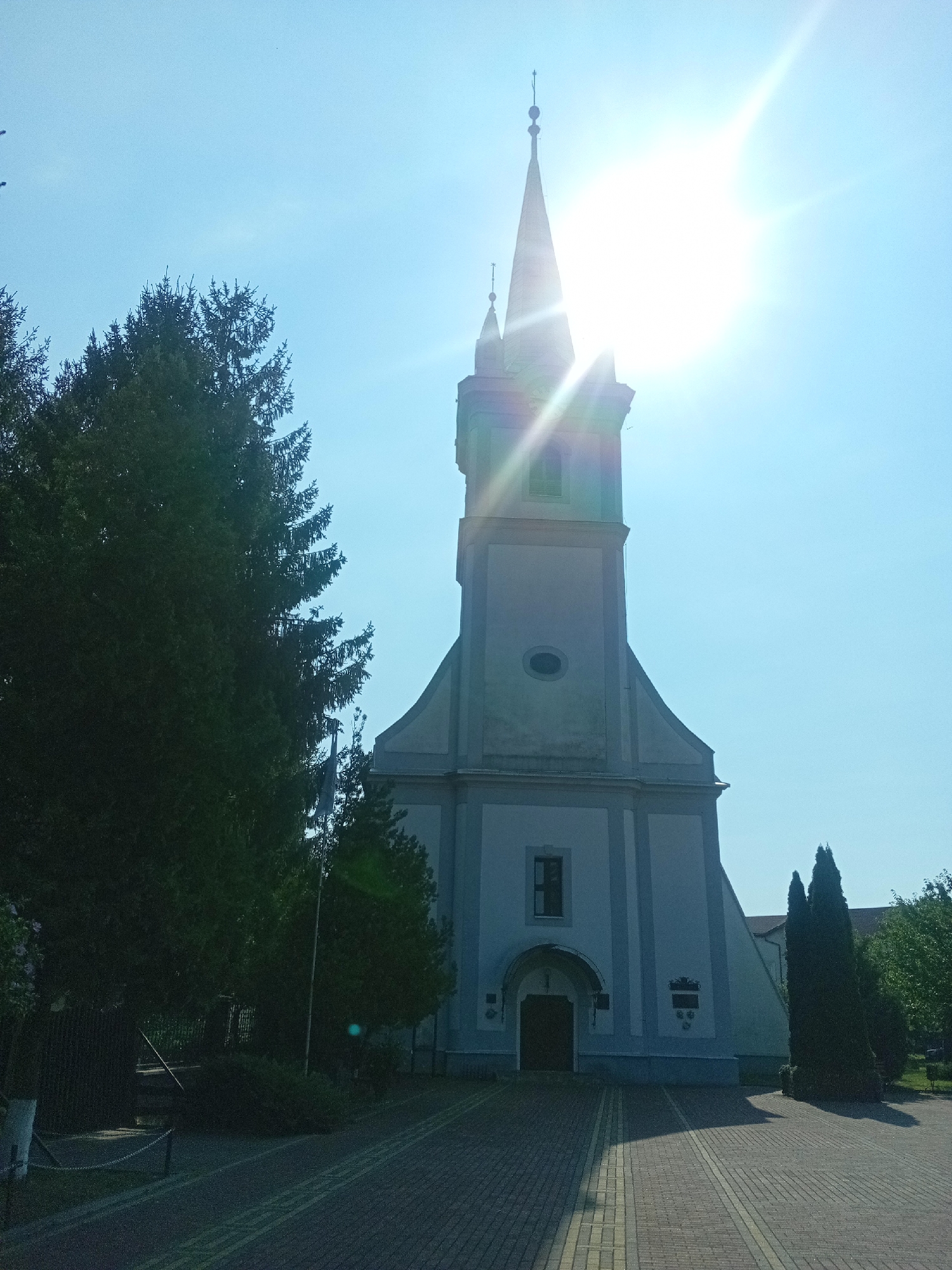 The image shows a light blue church with a tall, pointed spire against a bright, sunny sky. The sun is positioned behind the spire, creating a strong lens flare and glare across the upper portion of the image. To the left of the church, there is a large, dark green evergreen tree. In front of the church, a paved walkway made of bricks leads to the entrance. The walkway has yellow lines painted on it. To the right of the church, there are more trees and some fencing. The overall impression is a sunny day in front of a church. The location is identified as Tiachiv, Ukraine.