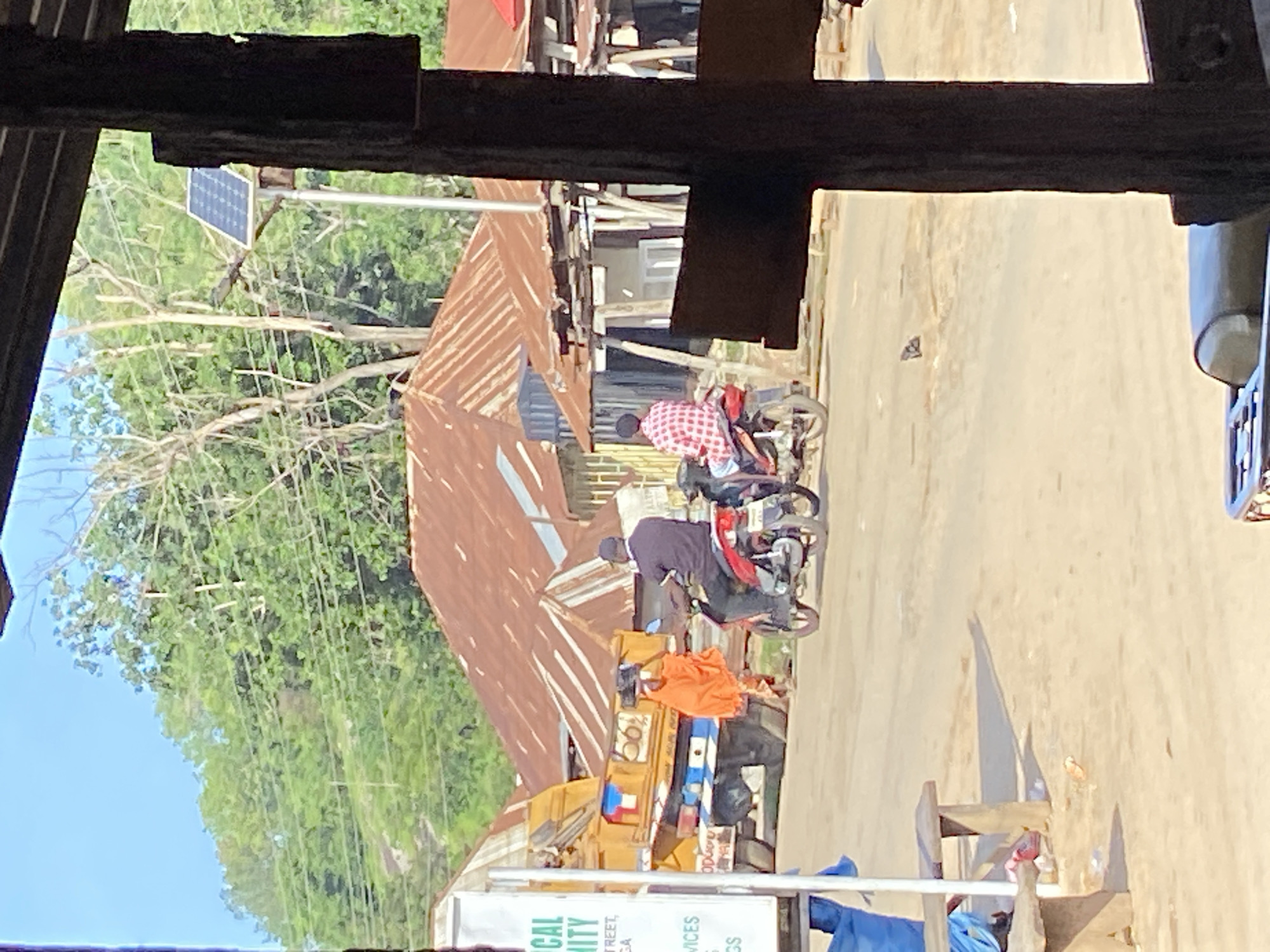 In Mararaba, Nigeria, a sunlit, dusty street scene unfolds. The foreground is partially obscured by a dark, weathered wooden post, suggesting a sheltered viewpoint like a veranda or shop. Beyond this, the street is populated with a few individuals and vehicles.
A yellow truck with markings, possibly a delivery or utility vehicle, is parked on the left side of the road. Behind it, a person in an orange garment walks while carrying an item on their head. Two men are riding motorcycles. The man in front, wearing a dark shirt, is on a red motorcycle. Behind him, a man in a red and white checkered shirt rides a red motorcycle.
The background features several buildings with rusty, corrugated iron roofs, indicative of the local architecture. Vegetation, including trees and foliage, covers the slopes of a hill in the distance. A solar panel is visible on a pole near the buildings, hinting at a blend of traditional and modern energy solutions. The overall impression is a typical day in a bustling Nigerian community, characterized by open-air commerce and the presence of motorbikes as common transportation. The ground appears to be unpaved, contributing to the dusty atmosphere.