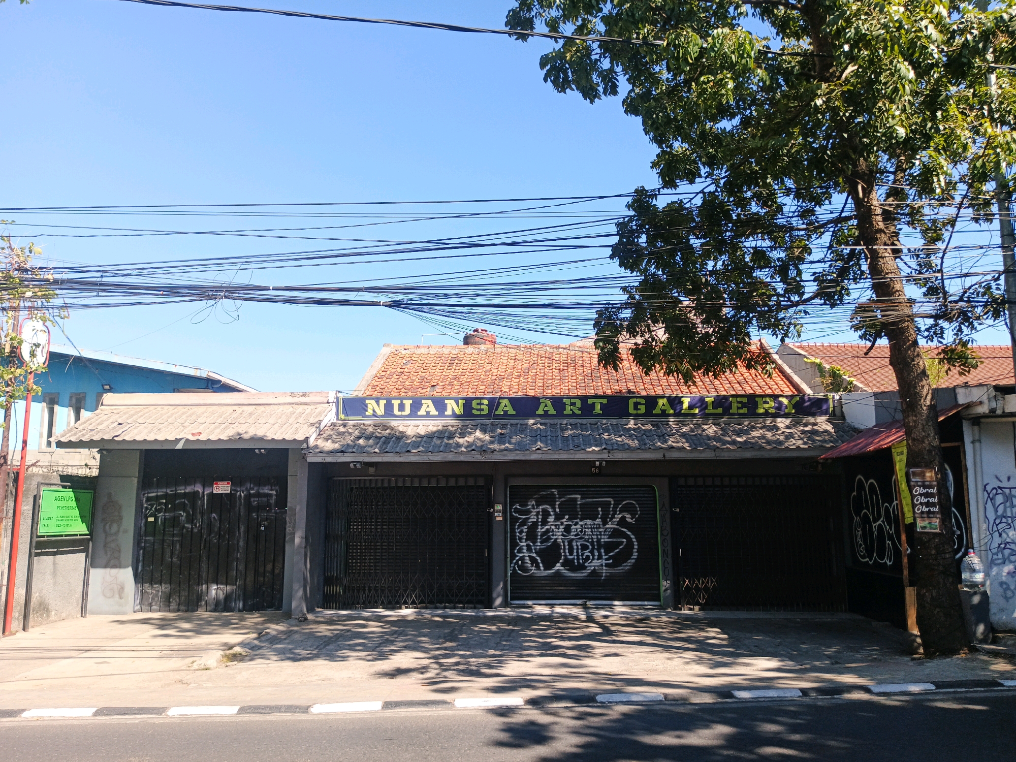 The image shows the exterior of a building with a sign that reads "NUANSA ART GALLERY". The building has a dark-colored, corrugated metal facade and is covered by a tiled awning. There are metal shutters on the windows and doors, some of which are covered in graffiti. A tree with green leaves stands to the right of the building. Overhead, a tangle of electrical wires crisscrosses the sky. The building appears to be located in a commercial or urban area. The building is located on Jalan Gatot Subroto, Bandung, Indonesia.