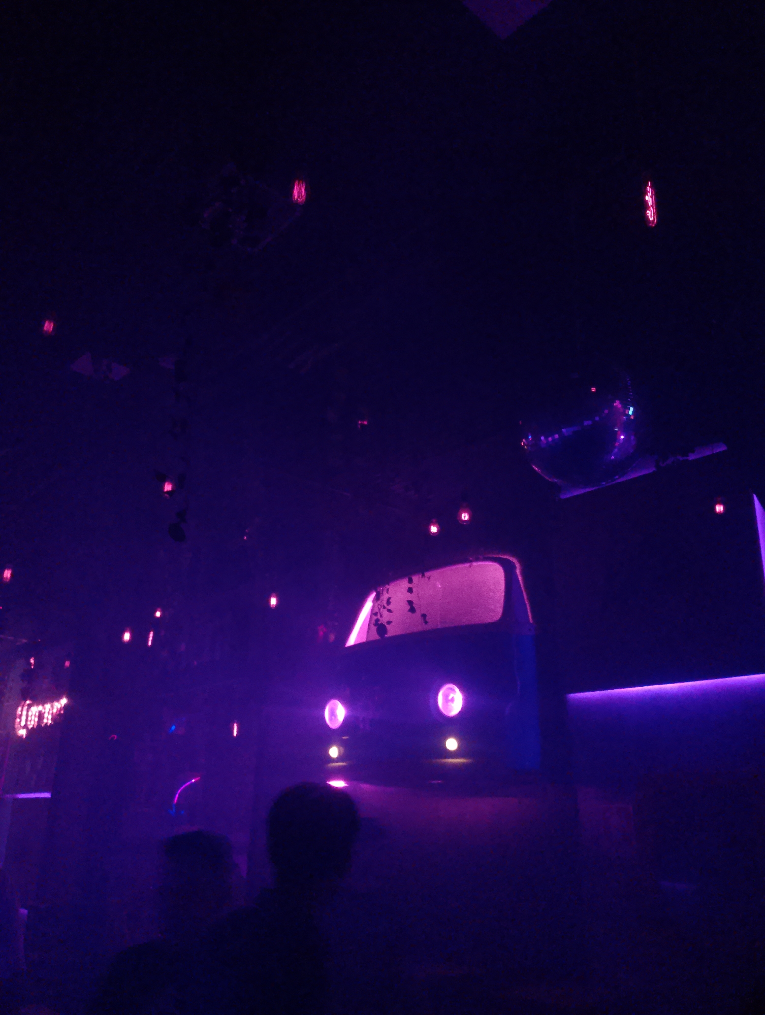 The image shows the interior of a nightclub or lounge in Porlamar, Venezuela, featuring purple lighting. In the foreground, the backs of two people's heads are visible. A vintage car, possibly a van, is elevated on a platform. The car's headlights are illuminated, and its windshield is faintly lit. Above and behind the car is a disco ball and other decorative lighting elements are scattered throughout the space.