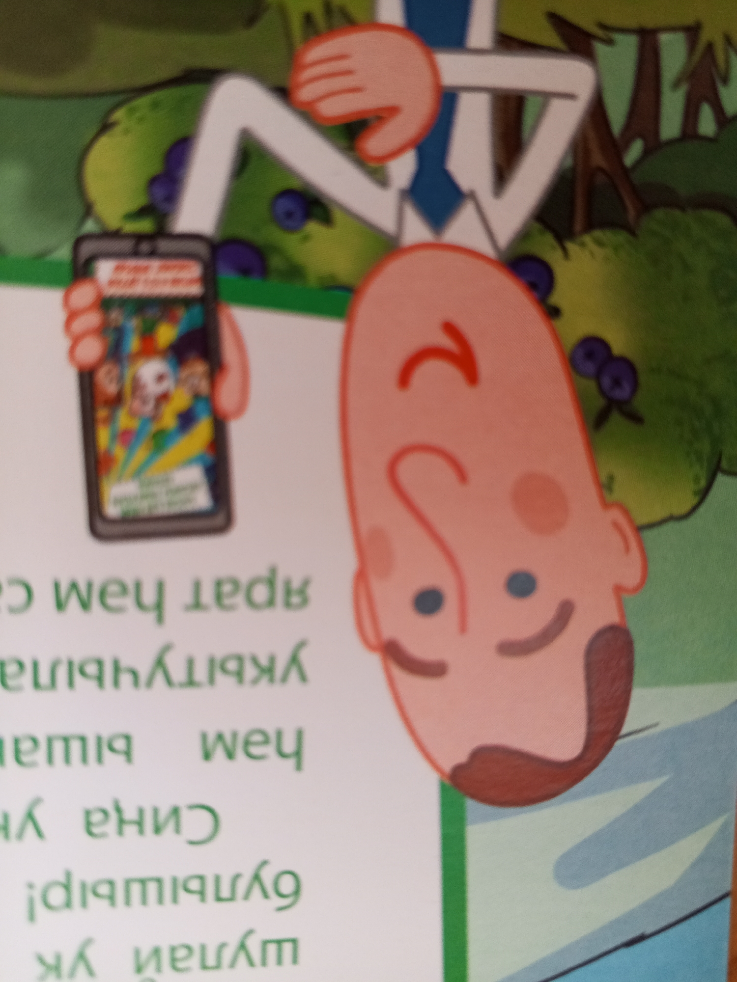 The image depicts a cartoon illustration of a person holding a smartphone. The smartphone screen displays a colorful image. The person's head is upside down, with features such as eyes, a nose, and a mouth visible. Another part of a person's arm and torso is visible to the left, also holding the phone. The background consists of greenery and trees. There is text present in the image, but it is blurry and difficult to read, and its location in Chistopol, Russia is not discernible from the image itself.