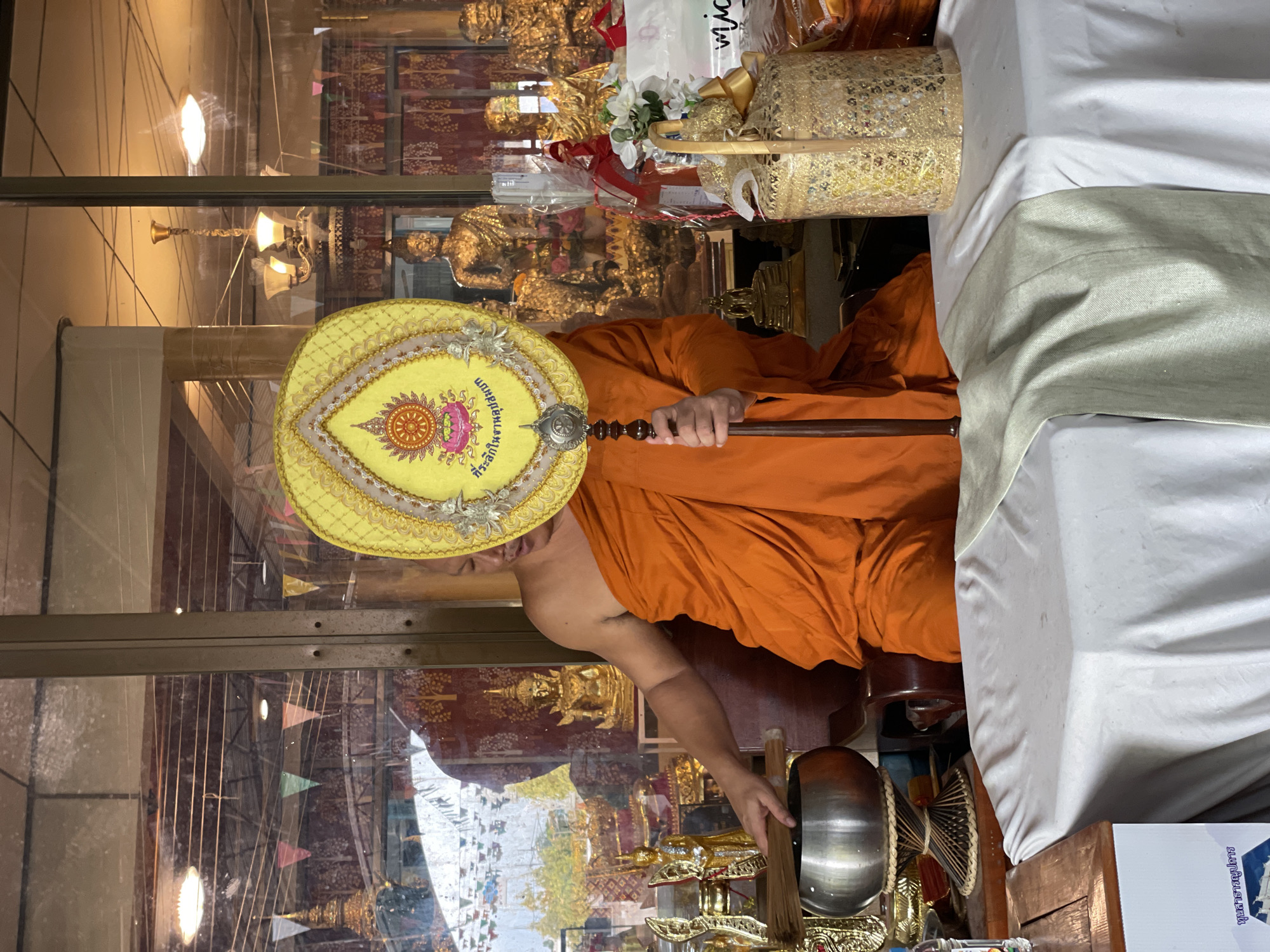 A monk, wearing an orange robe, stands behind a table covered with a white cloth in Tambon Na Chom Thian, Thailand. He holds a yellow fan with a circular emblem in front of his face, obscuring it. The fan has ornate gold trim and Thai writing. To the monk's left, a metal alms bowl rests on a wooden stand, with a bundle of sticks or brushes inside. The background appears to be an interior space with large windows revealing a shrine or temple area. Golden statues of Buddha are visible, along with decorative banners and a chandelier. On the table, to the right of the alms bowl, is a golden woven basket filled with flowers and ribbons, and various wrapped items.