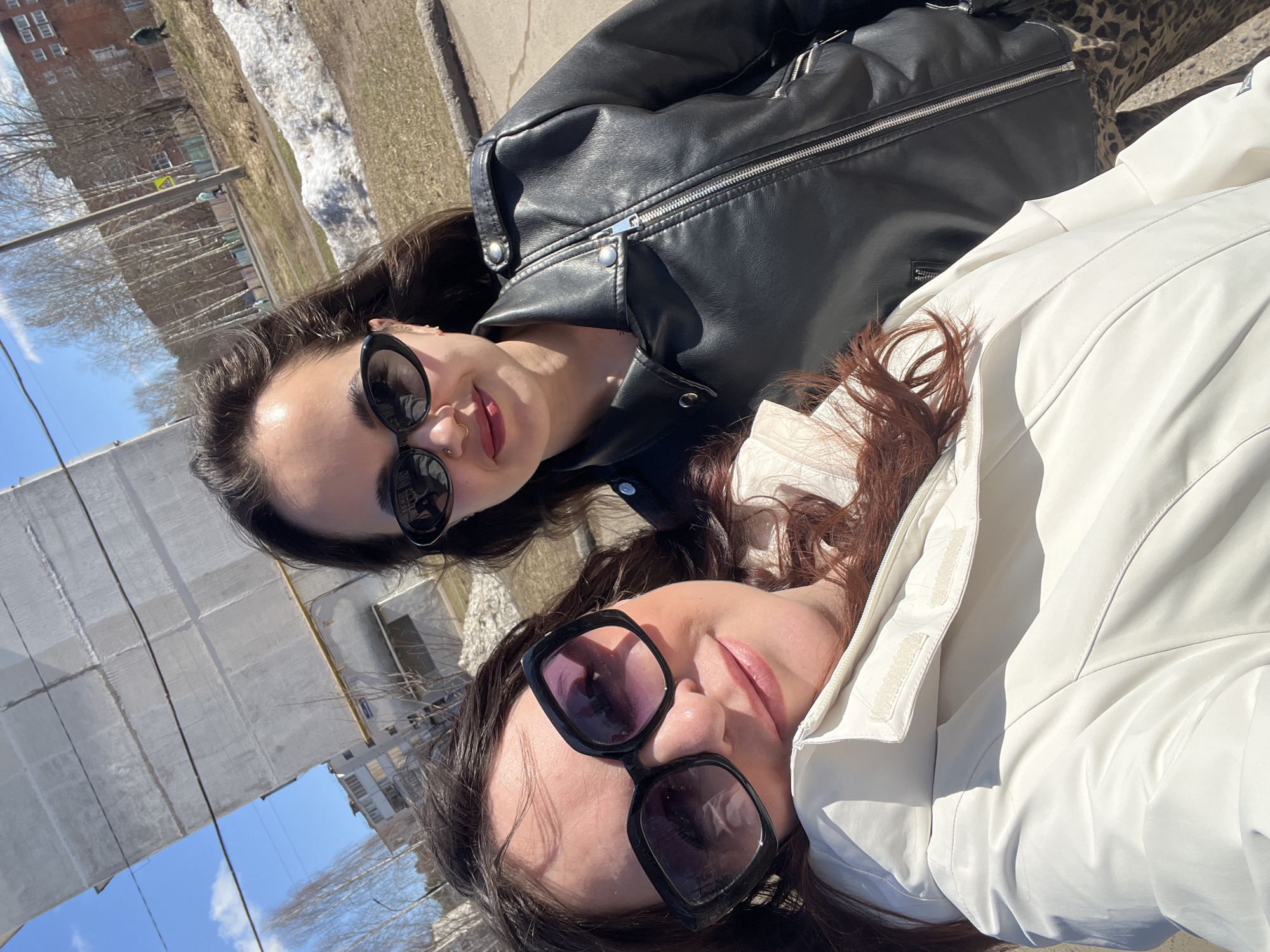 Two women are pictured in an outdoor setting, likely on a sidewalk in Noviy, Russia. The woman at the top wears a black leather jacket and dark sunglasses. The woman below her wears a white jacket and similarly styled dark sunglasses. Patches of snow can be seen nearby. A building with what appear to be apartments is visible. The sky is blue.