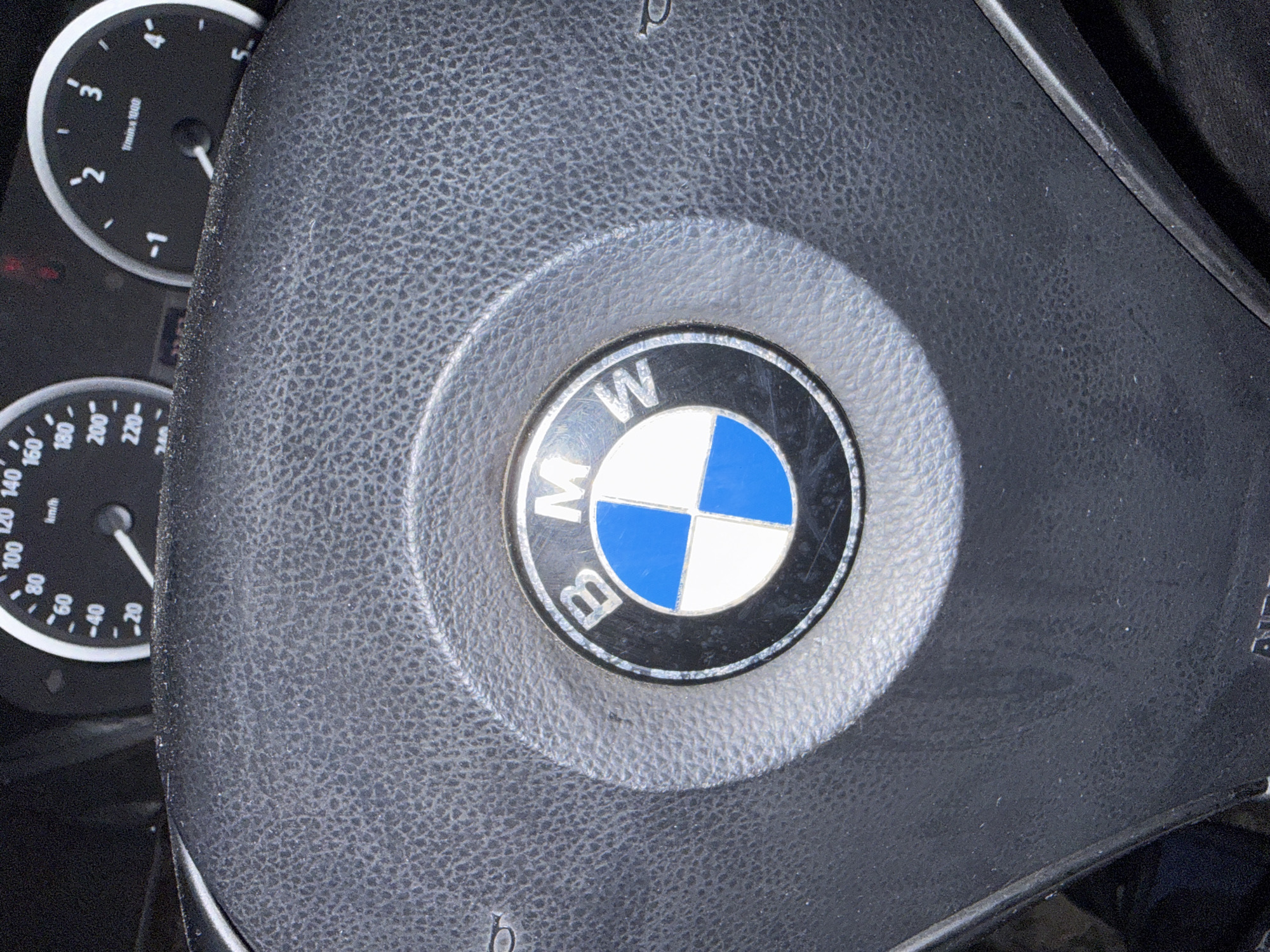 This image shows the interior of a vehicle, specifically the steering wheel with the BMW logo prominently displayed in the center. The logo is circular, featuring a black outer ring with "BMW" imprinted on it in white lettering. The inner portion is divided into quadrants of blue and white. The steering wheel itself is black and textured. To the left of the steering wheel, parts of the instrument panel are visible, including a tachometer and a speedometer. It is impossible to determine the location in Stara Zagora, Bulgaria or the presence of any people.