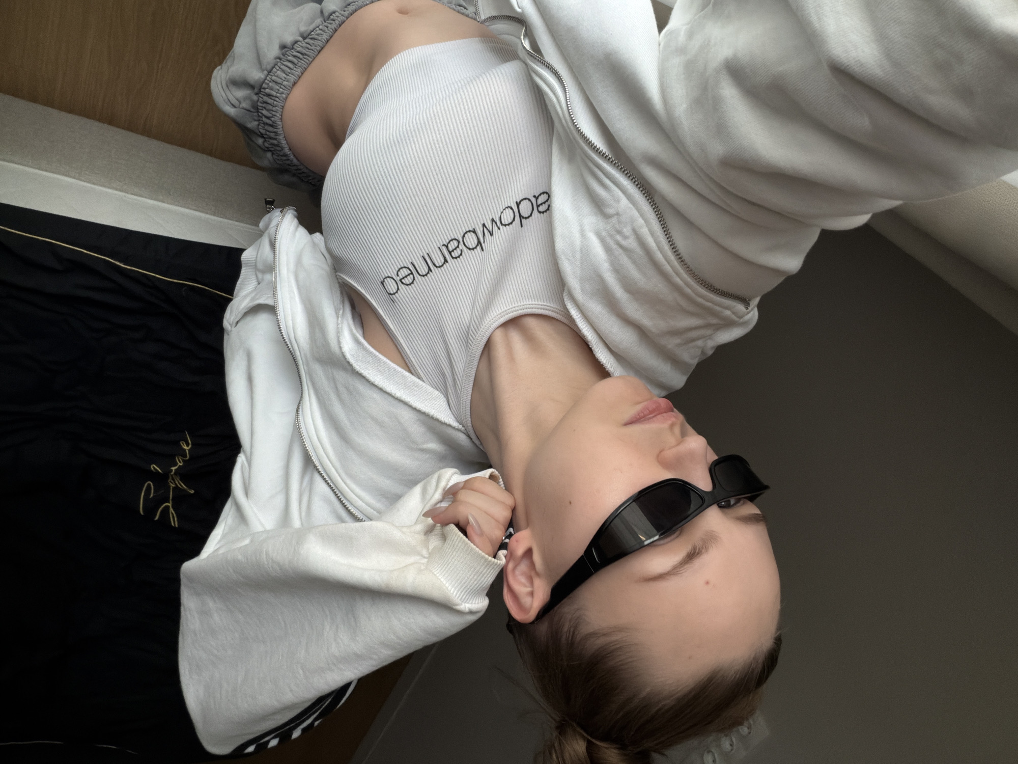 A female wearing sunglasses is angled on her back, against a neutral backdrop. She is wearing a white ribbed tank top with "dowbanned" written in black. A white zip-up hoodie is partially open, revealing the tank top. She also has on gray sweatpants. The setting includes parts of a bed and a wall, suggesting an indoor location, but there is no evidence of this image being in Moscow, Russia.