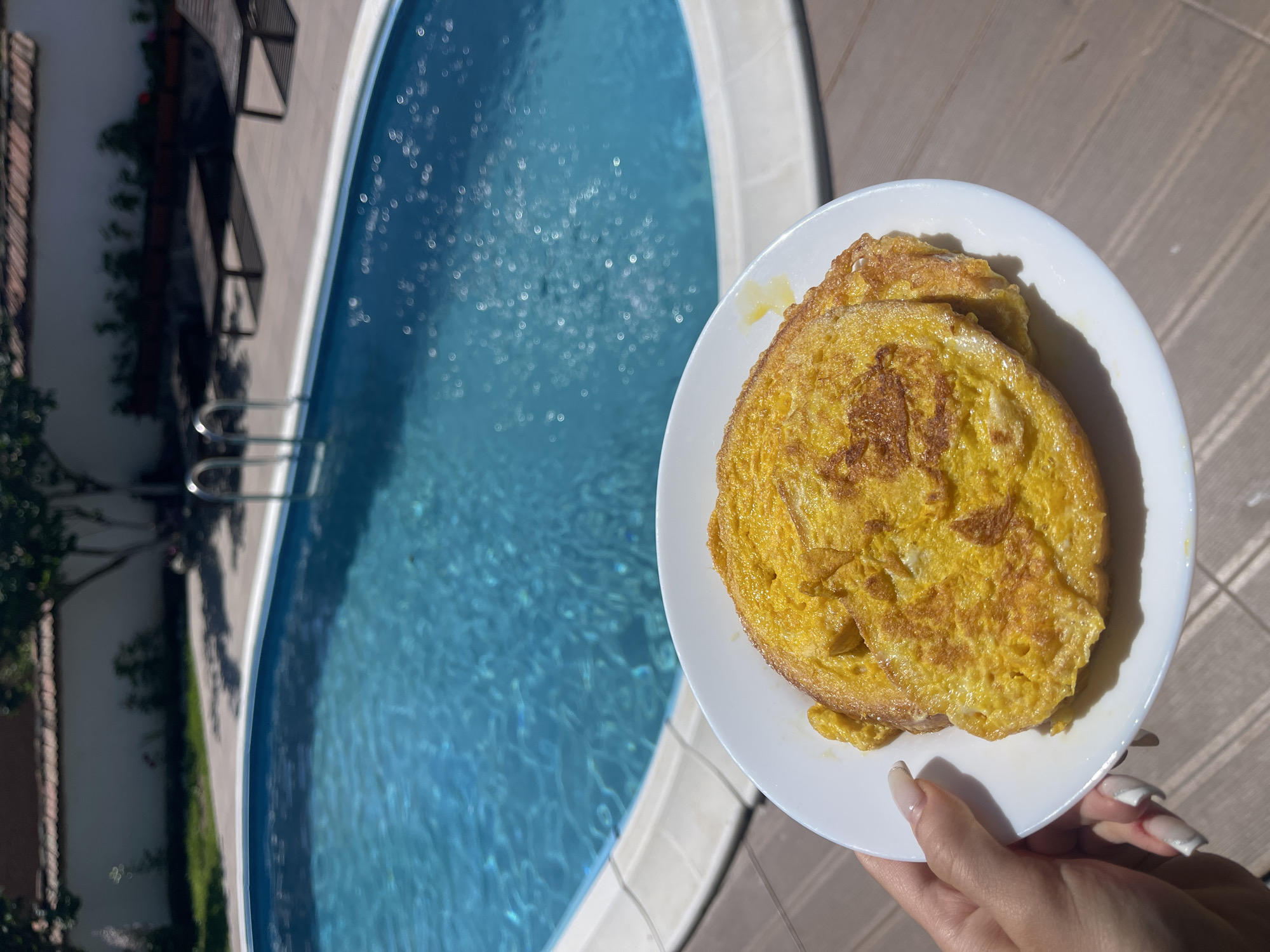 A white plate holds two pieces of French toast, a hand with manicured nails supports the plate, and a swimming pool is visible in the background. The swimming pool has blue water and lounge chairs are positioned around it. A wall and trees are behind the pool. This scene is likely located in Totleben, Bulgaria.