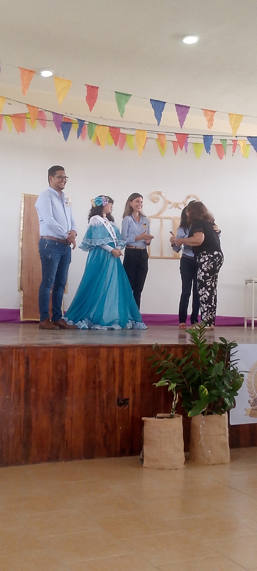 **Location:** Juan Griego, Venezuela.
**Scene:** An indoor stage setting.
**People:**
* A man standing on the left, wearing a light blue shirt and jeans.
* A woman in a blue dress and sash that reads "Reina de Fiestas" with a flower crown, indicating a queen or festival representative.
* Another woman, wearing a light blue button-up and dark pants.
* A woman in a black shirt and floral pants, hugging another woman.
**Objects:**
* A stage with a wooden front.
* Colorful triangular flags hung across the backdrop.
* Potted plants with burlap coverings.
* A wooden prop with decorative curls.
**Actions:**
* The man and woman stand on the left of the stage.
* One woman is hugging another woman, with the third woman standing next to them.
**Interactions:**
* An event is taking place on the stage where a woman is being thanked for her work in a festival, or congratulating her.
**Environment:**
* Bright interior.
* Minimal stage decorations.
* Tile floor.