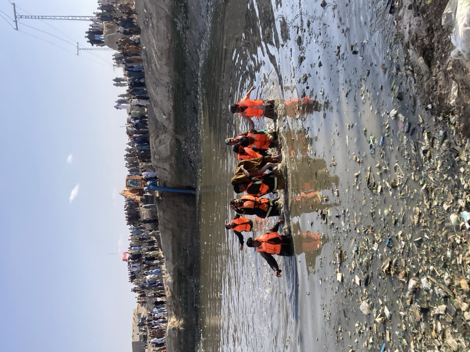 The image depicts a daylight scene in Quetta City Tehsil, Pakistan, showing a rescue operation within a shallow, heavily polluted body of water. In the foreground and mid-ground, six individuals, primarily wearing orange life vests over dark clothing, wade through murky, waste-filled water that reaches their waist or chest. They are actively supporting and moving a large, brown bovine animal, consistent with a cow or buffalo, which appears incapacitated. The water surface is densely covered with assorted debris, including plastic items and refuse.
On the elevated, dry earthen bank in the background, a large crowd of onlookers, predominantly male, observes the activity. An orange excavator with a black boom is positioned on this bank, with a large blue pipe or hose extending from it down into the water, suggesting a pumping operation. Utility poles and power lines are visible against a clear, light blue sky on the horizon.