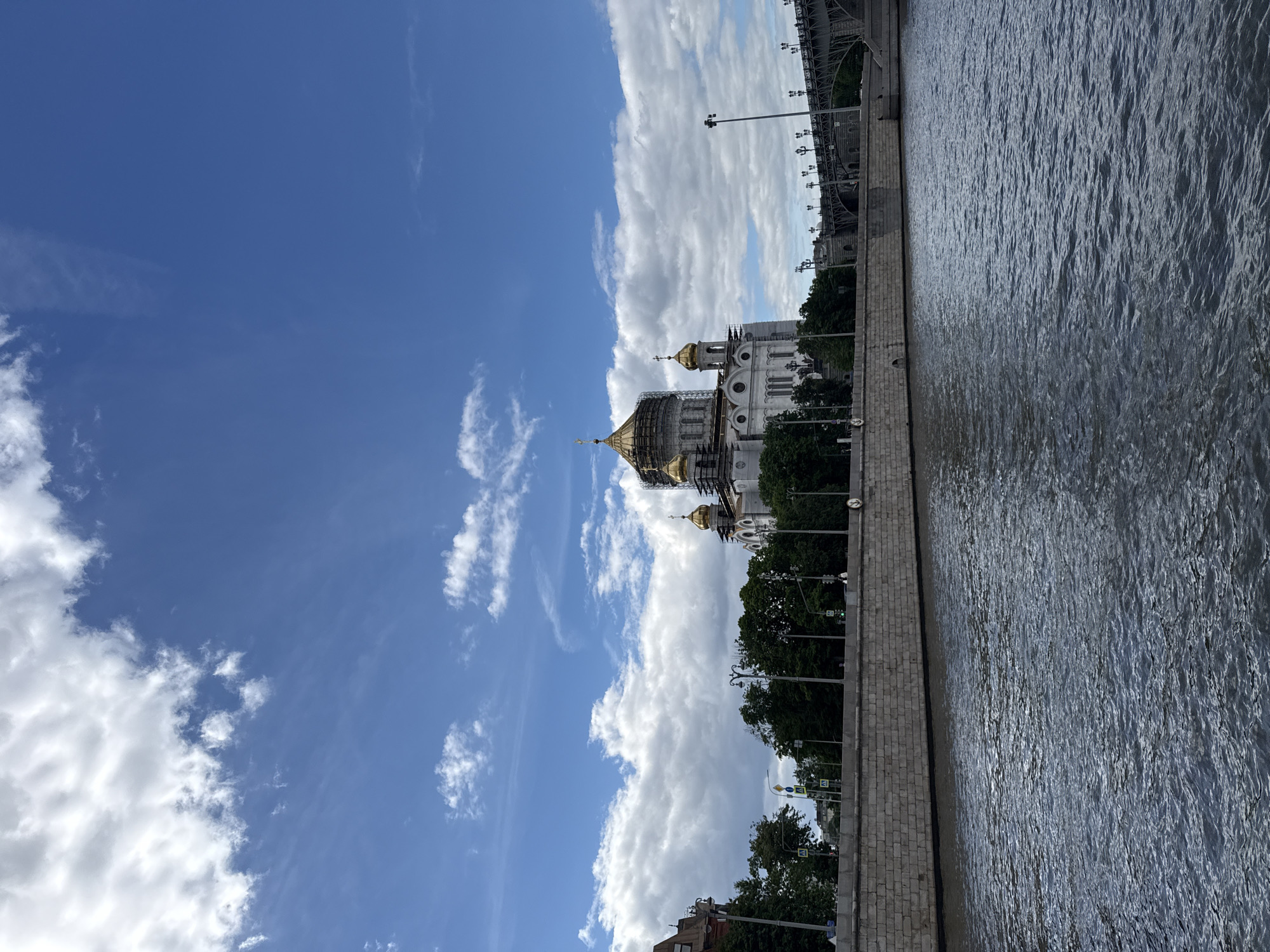 The image presents the Cathedral of Christ the Savior in Moscow, Russia, undergoing construction or renovation on its domes. A river runs along the right side of the frame, with a stone embankment separating it from the foreground. Trees line the embankment and partly obscure the view of the cathedral. Above, the sky is a mix of clear blue and scattered clouds. A portion of a bridge is visible in the top right corner. No people are visible in the image.