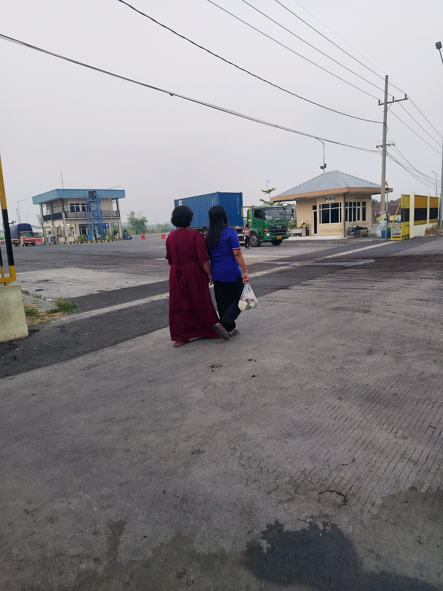 Two individuals are walking away from the camera on a paved surface in front of a small, open-air security post labeled "POS 9". The person on the left is wearing a long, dark red dress, and the person on the right is wearing a blue shirt and black pants, carrying a plastic bag with contents. Behind them, a green truck with a shipping container is visible, along with a multi-story building with blue accents in the background. The environment appears to be an industrial or port area, with a cloudy sky overhead. The location is identified as Kedung Sukodani, Indonesia.