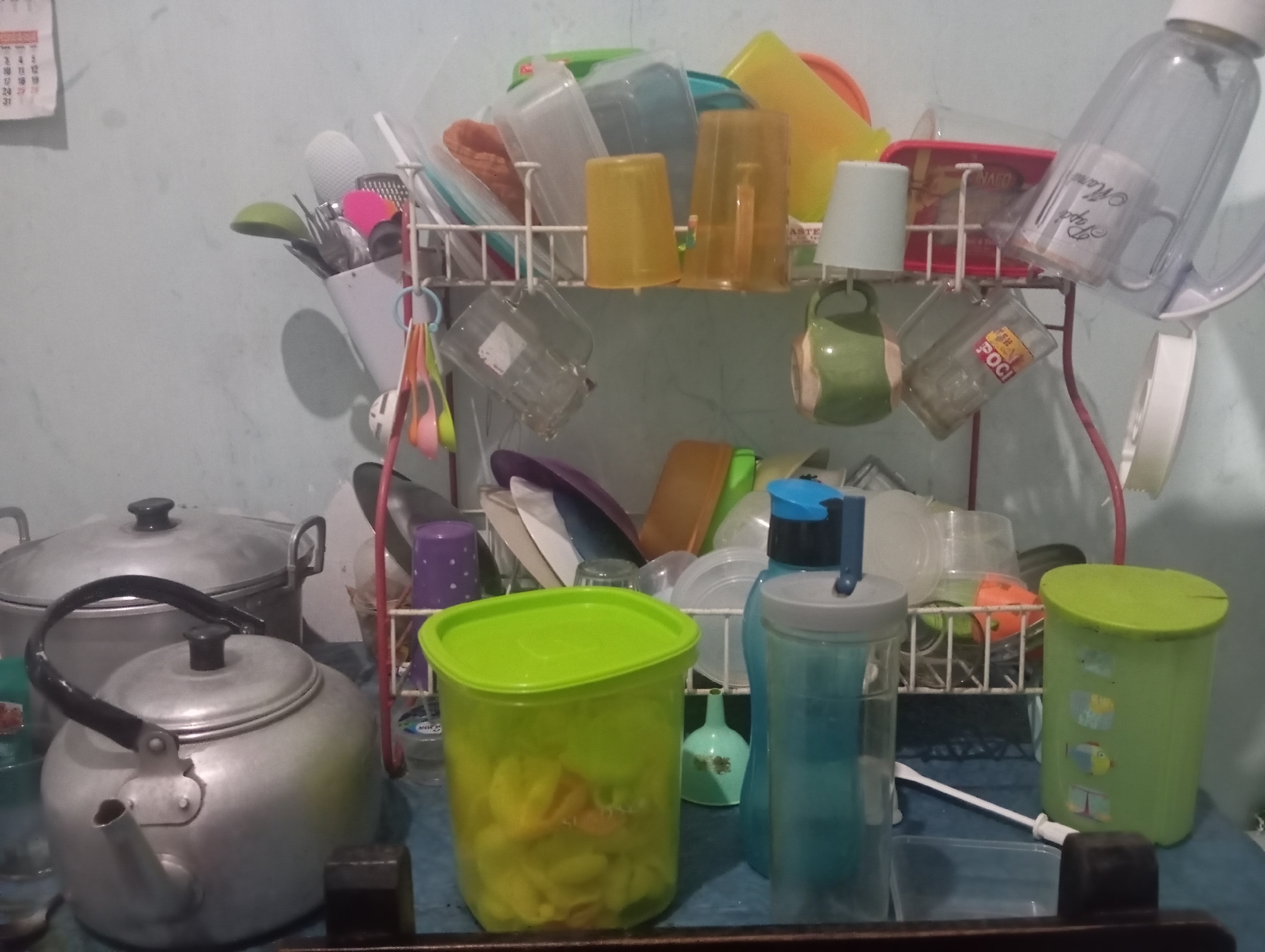 The image depicts a kitchen scene in Kutasari, Indonesia, with a dish rack filled with various kitchenware. The dish rack holds an assortment of plastic cups, containers, and other utensils. In the foreground, a silver teapot and a large aluminum pot are visible. To the right of the teapot, a green plastic container is filled with yellow food items, possibly snacks. Next to it, two blue plastic water bottles stand upright. In the background, the dish rack is laden with more containers, cups, and plates, with a white calendar hanging on the wall above. The overall impression is one of a functional, albeit cluttered, kitchen space.