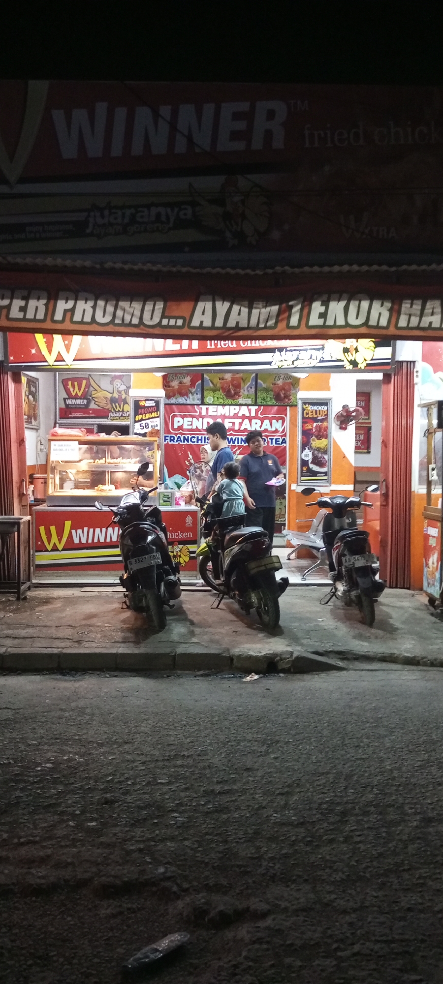 The image depicts a street-level view of a "Winner Fried Chicken" establishment, likely in Setu, Indonesia, during nighttime hours. The storefront is illuminated, revealing a red and orange color scheme. A prominent banner above the entrance reads "PER PROMO... AYAM TEKOR HA" with the "Winner" logo featuring a stylized "W" and a chicken graphic. Inside, a display case shows fried chicken.
Several people are visible outside the establishment, appearing to be customers or employees. Two individuals, an adult male and a younger child, are standing near the entrance, interacting with another adult male who is wearing a blue uniform and holding a plate. A third individual in a dark shirt is also present near the counter.
Three motorcycles are parked on the sidewalk in front of the store. The motorcycle on the left has license plate number B 3327 EBU. The motorcycle on the right has license plate number AB 3474. The ground in front of the store is paved, and a sidewalk area is present. The surrounding environment appears to be a street with a dimly lit background.