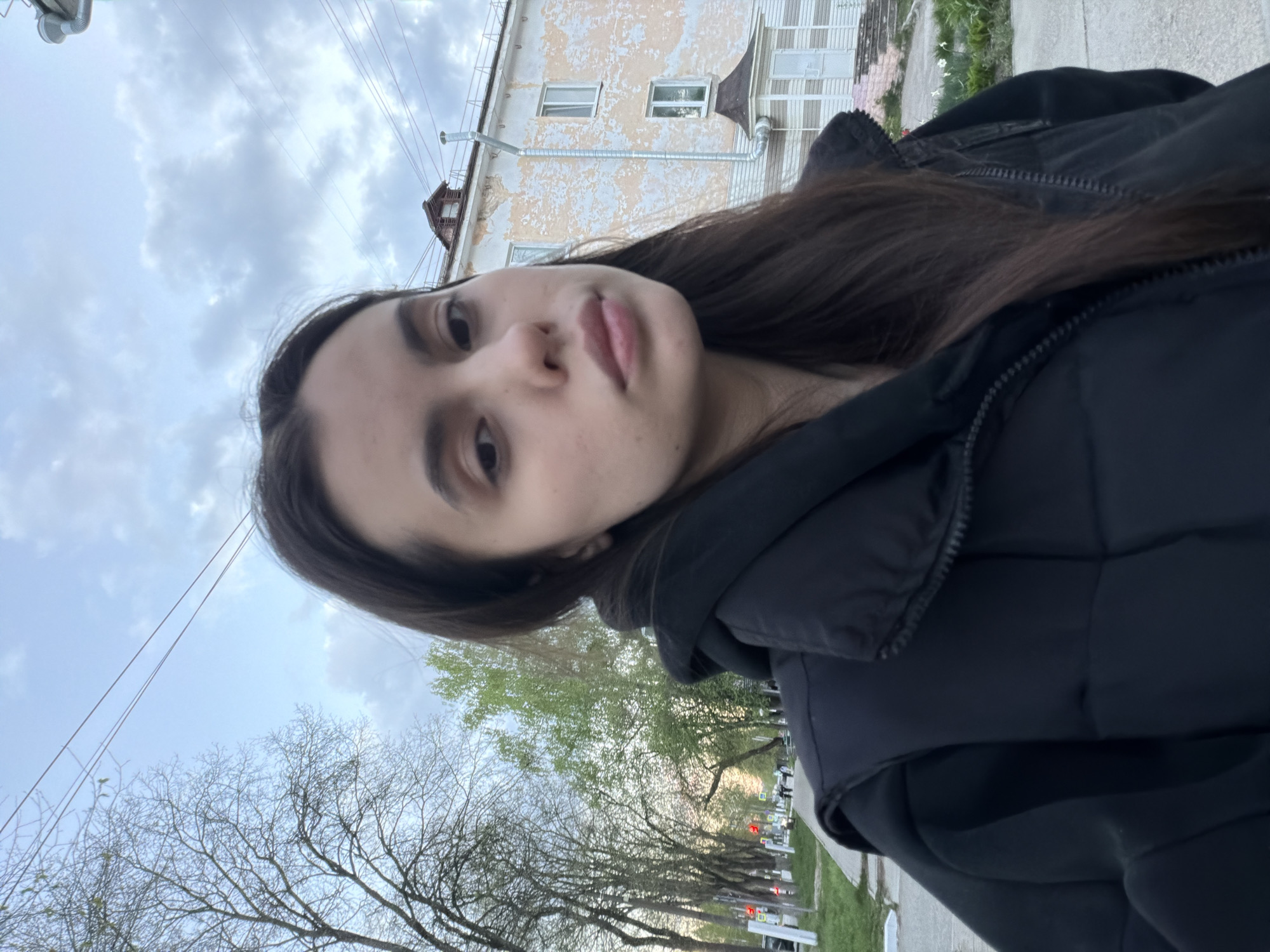 A young woman is taking a selfie in Chaikovsky, Russia. She is wearing a black puffer jacket. Behind her is a building with peeling paint and visible electrical wires. Trees line the street in the background. The sky is overcast.