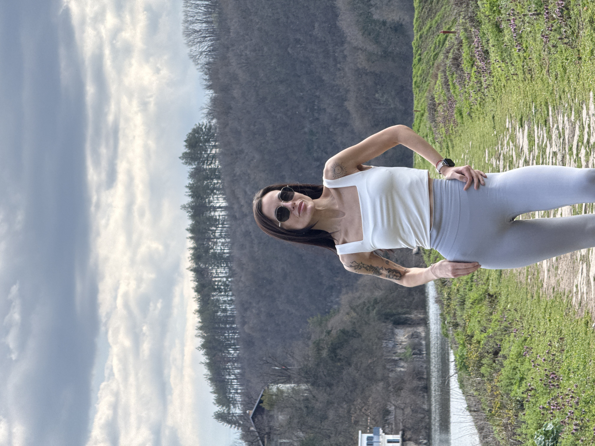 A forensic examination of the image reveals a female subject standing on a dirt path in an outdoor environment, identified as Kaylaka Park (Park "Kailuka") in Pleven, Bulgaria. The subject, a woman with long dark hair, wears dark sunglasses, a white square-neck crop top, and light grey athletic leggings. Tattoos are visible on both upper arms: a detailed floral or vine pattern on the left, and a sun and crescent moon design on the right shoulder. A smartwatch with a pink and white band is present on her left wrist. She stands frontally, with her hands resting on or near her hips, exhibiting a slight smirk.
The immediate foreground features an unpaved path composed of dirt and scattered light-colored stones, flanked by green grass dotted with small clusters of purple flowers. To the left of the path, a calm body of water, consistent with a reservoir or dam lake, extends into the midground. The far bank of the water is steep and rocky, supporting a white building with blue window frames. Further back on a rising hill, a larger, light-colored building with a dark roof is visible, from which a flagpole flies a red, white, and green striped flag, indicative of the national flag of Bulgaria. The background consists of extensive hills covered predominantly by bare deciduous trees, suggesting late winter or early spring. A clear line of coniferous trees crowns the highest visible ridge. The sky is overcast with multiple layers of grey and white clouds.
