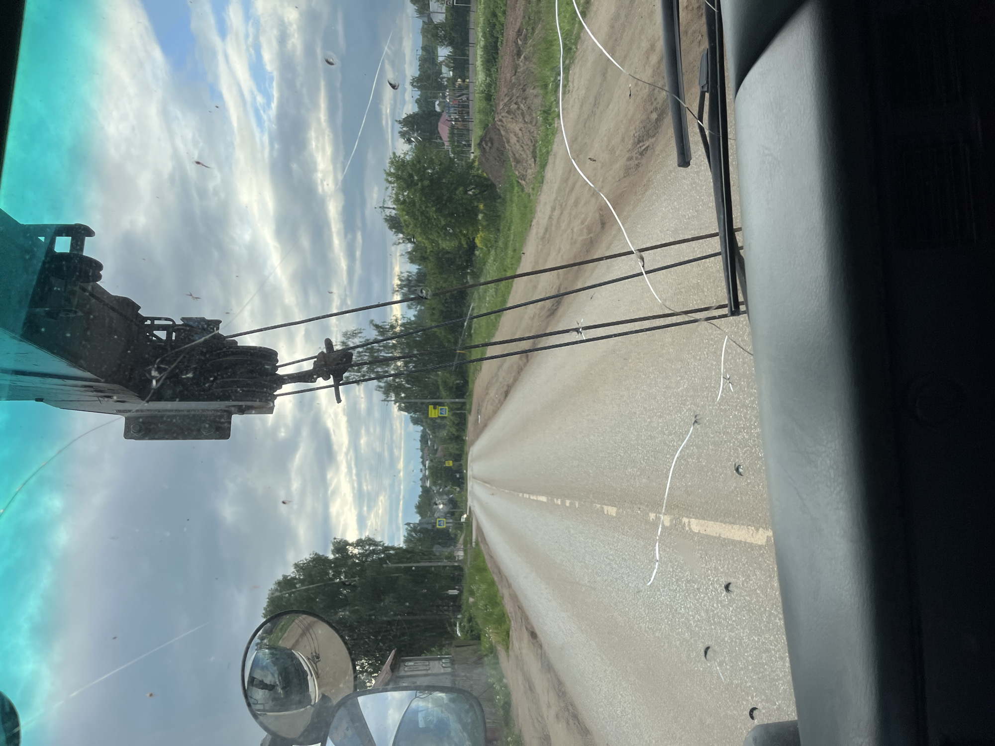The image depicts a forward-facing view from inside the cab of a heavy-duty vehicle, likely a crane truck, positioned on an asphalt road in Фоки, Russia, under an overcast sky. The vehicle's windshield is heavily cracked, particularly across the bottom half, and appears dirty with scattered debris and insects. A teal-tinted sun visor is visible across the top of the windshield. On the left, a side mirror reflects a two-story wooden building and surrounding trees. On the right, a significant portion of a crane boom, including its pulley system and cables, is visible through the windshield, extending downwards from the upper right.
The road ahead is a two-lane paved surface with a faded white center line, partially covered in dirt and sand, suggesting unpaved shoulders. Along the left side of the road, dense green foliage and trees line the view, with a multi-story building set back from the road. Utility poles and wires are also present. On the right side of the road, the immediate area consists of a dirt path and grassy terrain, leading to several large piles of brown earth or sand. Further in the distance on the right, a red-roofed building and a playground with colorful structures are partially visible amidst more trees. Two yellow triangular road signs, depicting children or pedestrians, are visible in the mid-distance on both sides of the road. The sky is predominantly cloudy, with patches of lighter sky visible. No individuals are discernible in the scene. The vehicle's black dashboard and a windshield wiper are visible at the bottom of the frame.