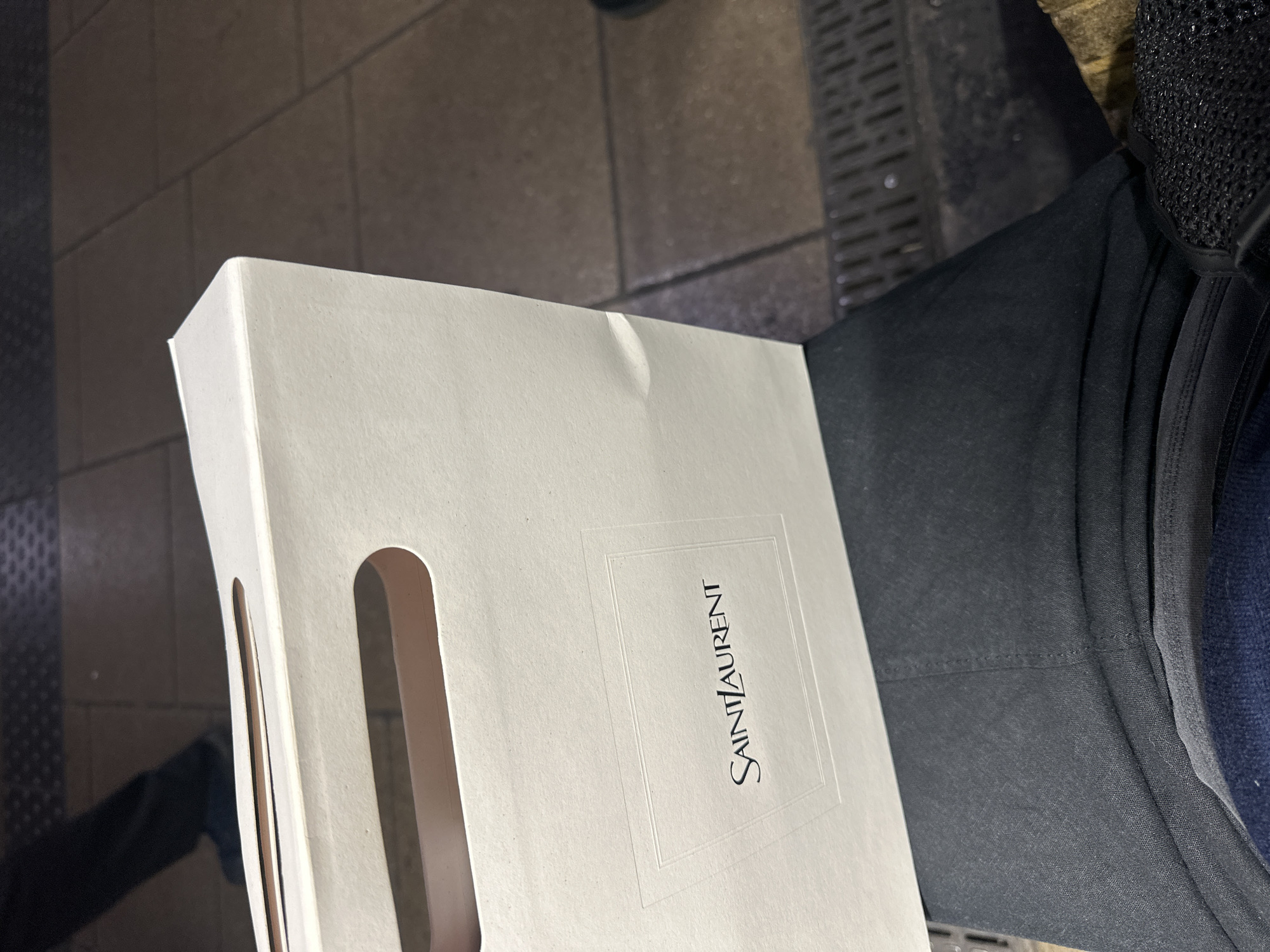 An off-white Saint Laurent shopping bag, featuring an embossed "SAINT LAURENT" logo within a rectangular frame, is positioned on the lap of an individual. The bag displays die-cut handles with a brown interior lining visible through the openings. The individual is wearing dark-colored trousers and a dark blue garment beneath. A black, mesh-textured object is partially visible on the lower right side of the individual's lap. The scene is set in a public transport environment, identifiable as a station platform in London, United Kingdom. The flooring consists of worn brown and grey square tiles, with a dark metal drainage grate visible to the right. In the blurred background, the lower legs and dark footwear of another person are visible moving across a textured platform edge.