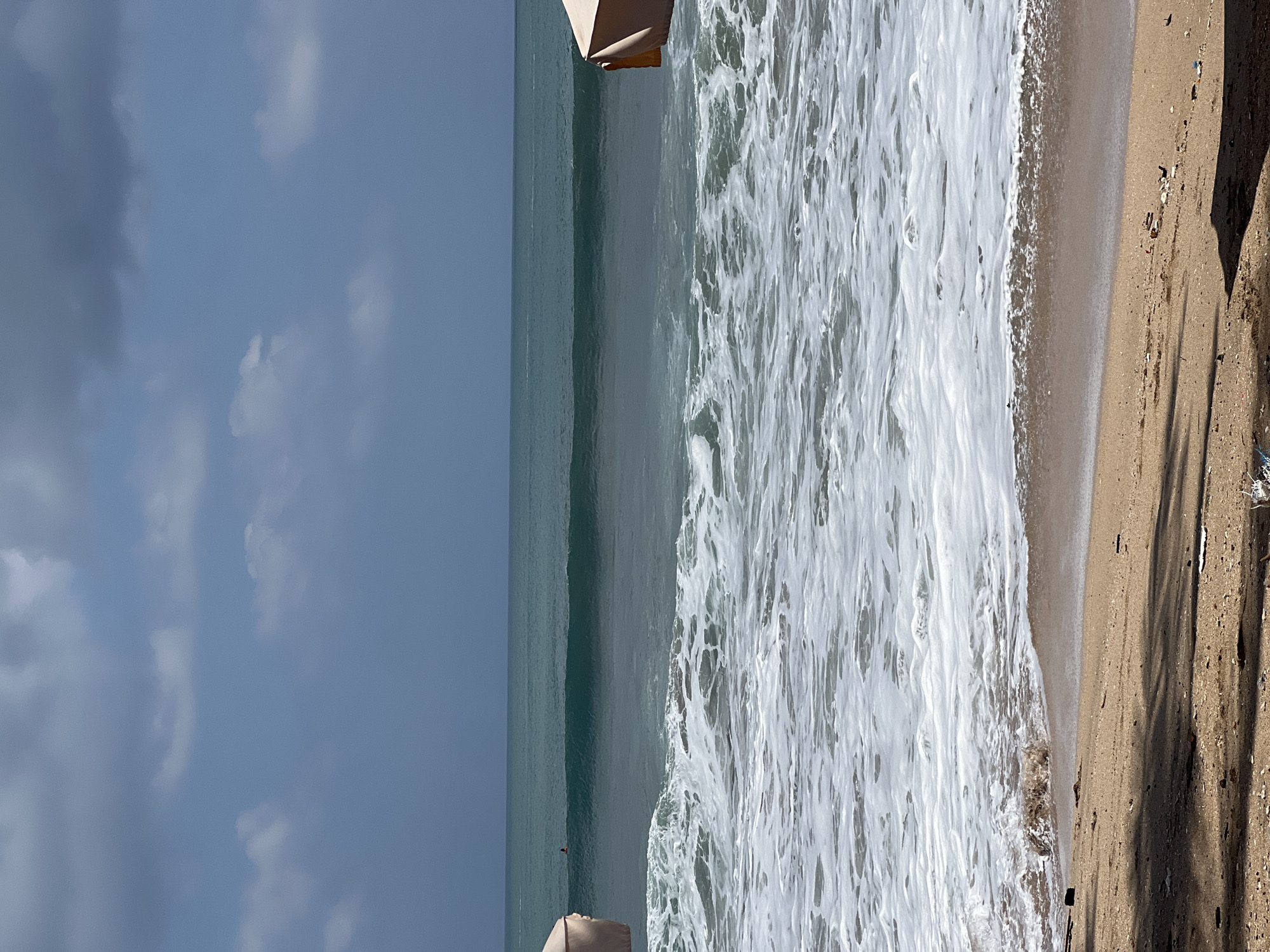 The image captures a scenic view of Kuta Beach in Bali, Indonesia. The foreground features a sandy beach with scattered debris. In the mid-ground, the ocean displays rolling waves with white foam crashing onto the shore. A surfer is visible in the distance, riding a wave. The background consists of the expansive sea and a partly cloudy sky. Two beach umbrellas are partially visible on the left and right sides of the frame.