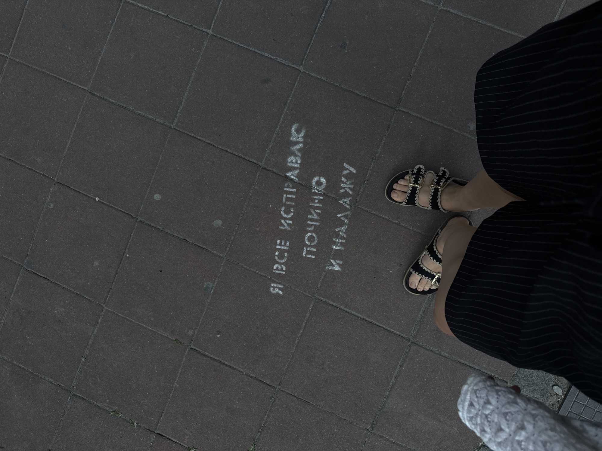 The image shows a top-down view of a person's legs and feet standing on a tiled sidewalk in Belgrade, Serbia. The text "Я ВСЕ ИСПРАВЛЮ ПОЧИНЮ И НАЛАЖУ" (I will fix everything, I will start and I will fix it) is stenciled in white on the dark grey tiles. The person is wearing black striped shorts and black sandals with gold buckles. Part of a white lace garment is visible to the left of the frame. The setting appears to be an outdoor urban environment.
