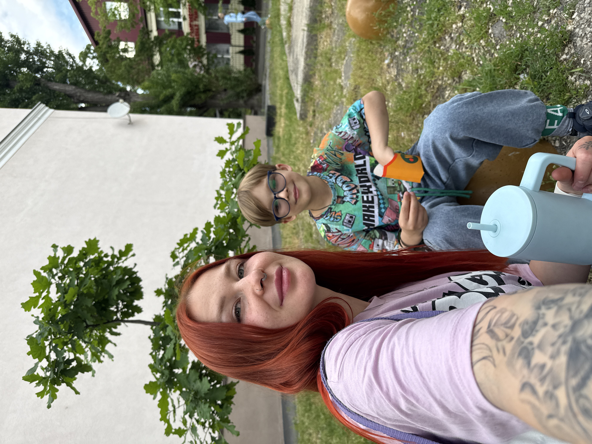The photograph captures a young woman with long red hair, wearing a light purple t-shirt, seated outside with a young boy who has glasses. The boy is wearing a colorful t-shirt and gray pants. The woman has tattoos on her arm. They appear to be outside on grass next to a white building with green trees. A building with dark red walls and white window frames is visible in the background. A pale blue container with a straw is visible in the foreground. There is no indication that this is Rostov-on-Don, Russia.