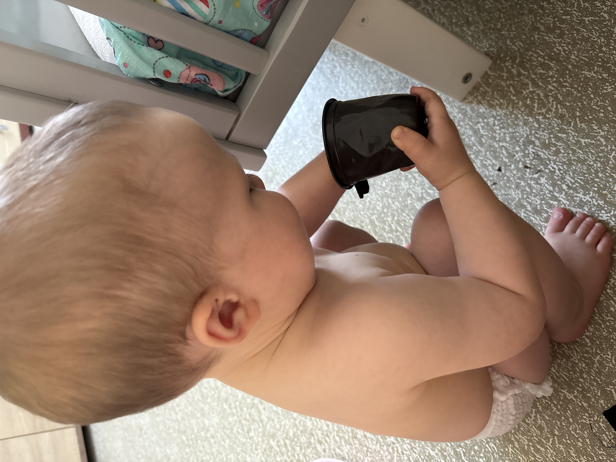A close-up, overhead shot captures a baby sitting on a patterned floor, possibly a rug or carpet, next to a light-colored furniture piece. The baby is turned away from the camera, looking towards the furniture. The baby is wearing only a diaper with ruffled edges. The baby's skin is fair, and their hair is light brown.
In the baby's right hand, they are holding a small, dark-colored cup or container, possibly a disposable cup, with a finger inserted into it. The baby's left arm is bent, resting on their lap. The baby's legs are bent and crossed. The baby's feet are visible at the bottom right of the frame, with toes clearly defined.
Underneath the furniture, some colorful baby bedding or fabric is visible, featuring pastel colors and patterns of hearts and circles. The furniture itself appears to be made of light-colored wood or painted material, with a visible screw on its leg. The lighting suggests it is daytime, with natural light illuminating the scene. The background floor has a textured, speckled pattern. The overall impression is of a candid moment of a baby playing with an everyday object in a domestic setting.
