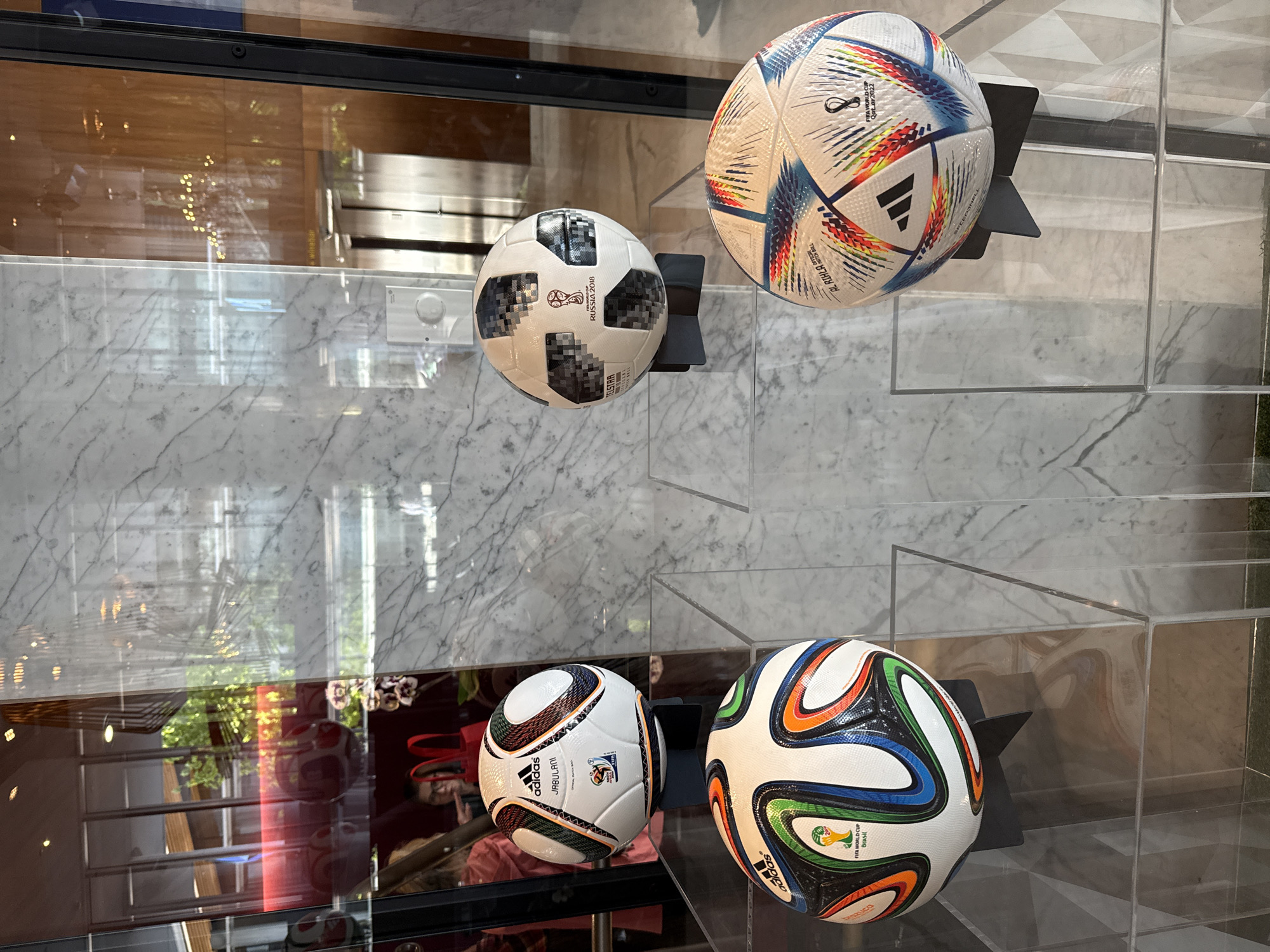 The image shows a display of four soccer balls, each from a different FIFA World Cup tournament. The balls are placed on black stands inside a clear glass display case with a marble-patterned background.
From top left to bottom right, the balls are:
1. Russia 2018 - Telstar
2. Qatar 2022 - Al Rihla
3. South Africa 2010 - Jabulani
4. Brazil 2014 - Brazuca
The display case appears to be indoors, based on the surrounding reflections, possibly in a retail or public space. There is a complex reflection in the glass display case, obscuring the exact details of the surroundings. The picture was taken in Vancouver, Canada.
