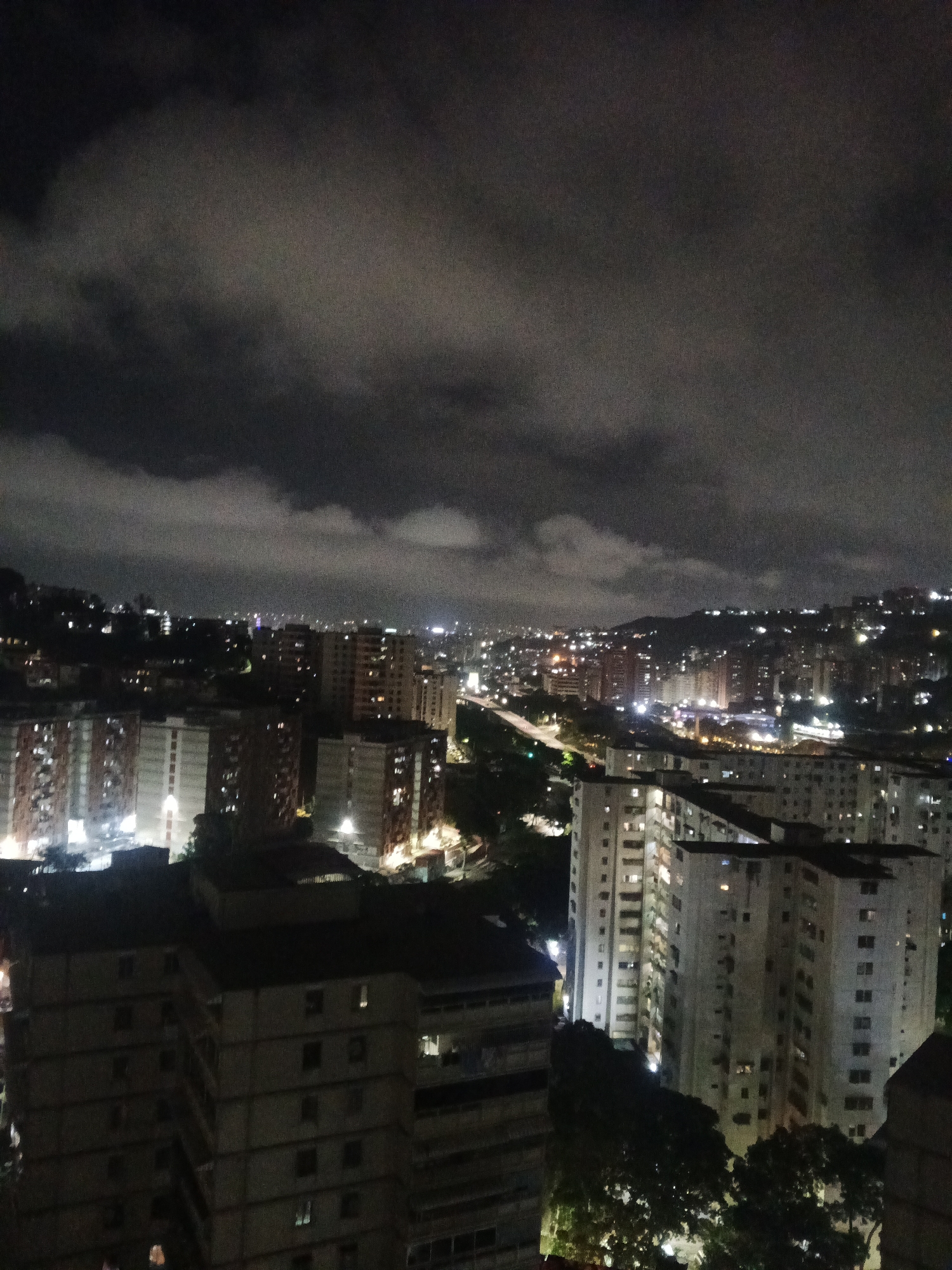 The image depicts an urban cityscape at night, likely taken from an elevated position, as indicated by the view of multiple buildings and a horizon of distant lights. The location is Caracas, Venezuela, as suggested by the dense arrangement of high-rise residential buildings, a common architectural style in the city, and the surrounding hilly terrain.
The foreground is dominated by several large, multi-story apartment buildings, typical of Caracas residential areas. These buildings are dimly lit, with lights visible in some windows, indicating occupancy. The architectural style is functional and lacks ornate detailing, reflecting a utilitarian approach to urban housing.
In the middle ground, a network of roads and highways is visible, illuminated by streetlights and vehicle headlights. This network indicates the city's transportation infrastructure and its role in connecting various parts of the urban area.
The background reveals a sprawling urban landscape extending into the distance. The horizon is dotted with clusters of lights, representing the city's outer limits and the surrounding hills. Above the city, a heavy cloud cover obscures the sky, creating a dark and moody atmosphere.
Overall, the image portrays a typical urban scene in Caracas, characterized by dense residential development, active transportation networks, and a cloudy night sky. The image does not feature any people, visible actions, or interactions. The focus is on the urban environment and its physical characteristics.