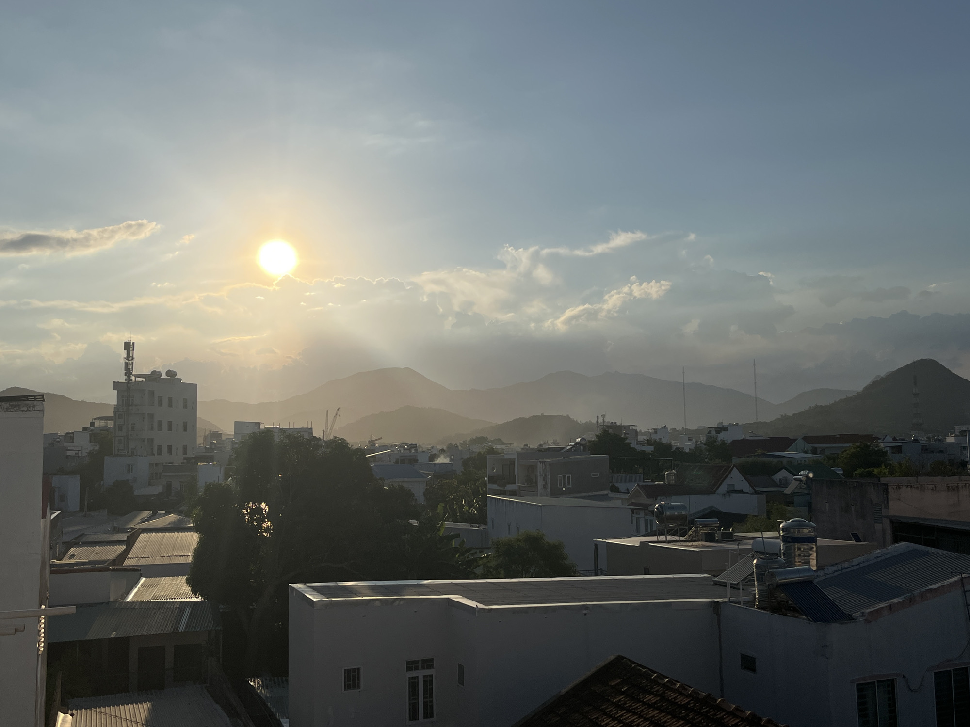 This image is a landscape photograph taken from a high vantage point, likely a rooftop, overlooking the city of Bac Nha Trang Ward, Vietnam, during what appears to be sunset. The sun, a bright, glowing orb, is visible in the upper left quadrant of the sky, casting long rays of light through the scattered clouds. The sky transitions from a pale blue at the top to a warmer, golden hue near the horizon, where it meets a range of distant, hazy mountains.
The city itself is densely packed with buildings of various sizes and architectural styles, predominantly low-rise structures with flat or corrugated metal roofs. Several taller buildings, including what appears to be an apartment or office building with multiple windows and an antenna on its roof, are scattered throughout the urban landscape. Trees are interspersed among the buildings, some casting dark shadows in the fading light.
In the foreground, the edge of another building's roof is visible, providing a framing element for the scene. The overall impression is one of a bustling urban environment bathed in the warm, atmospheric light of the setting sun, with a backdrop of natural mountain scenery. There are no people or discernible actions depicted in the image. The focus is on the panoramic view of the city and its environment.