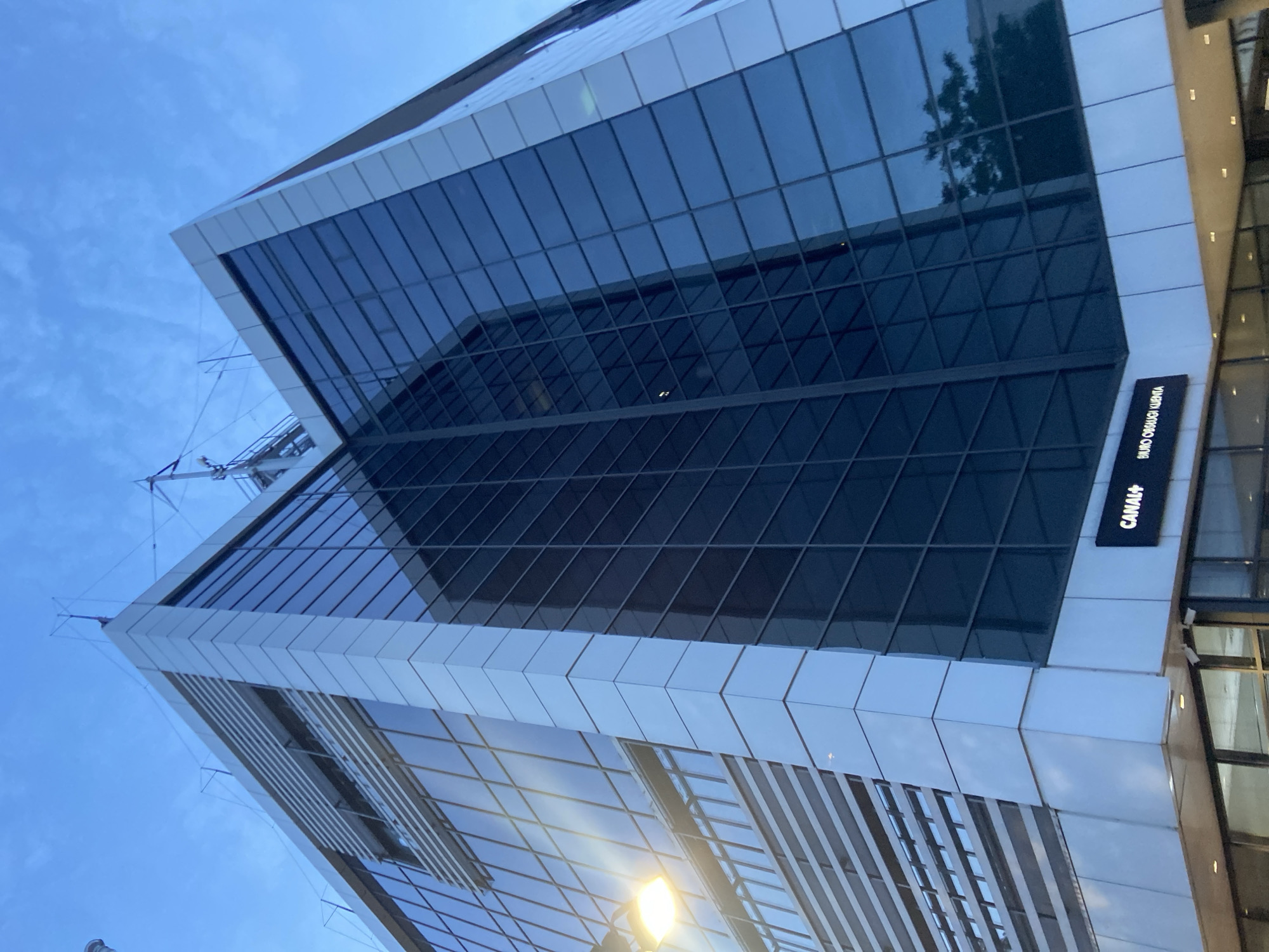 The image shows the Canal+ building in Warsaw, Poland, at dusk. The modern, multi-story building features a facade of glass and metal. The glass panels are reflective, mirroring the sky and adjacent structures. A streetlamp is visible on the left side of the image, casting a warm light. The sky is a deep blue, with a few wispy clouds. The building's exterior is clean and well-maintained. A sign displaying "CANAL+" is prominently displayed on the facade. The overall impression is one of a contemporary urban landscape.