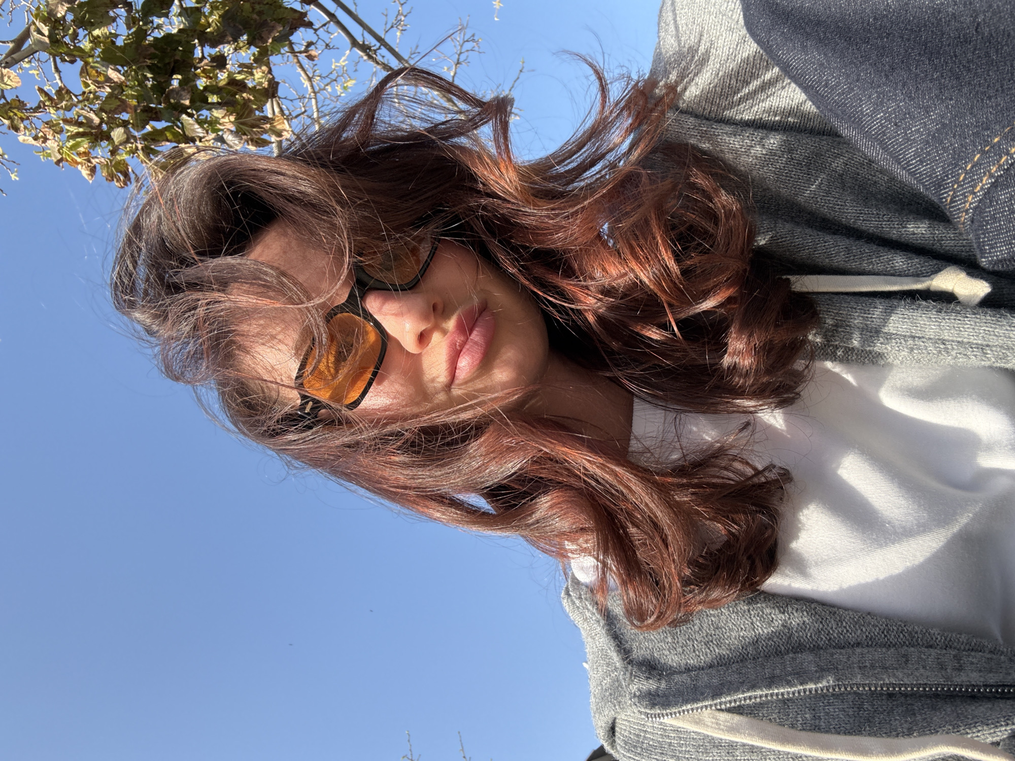 A woman is photographed from a low angle, looking down towards the camera, with blue sky and sparse tree foliage visible in the background. The woman is wearing tinted sunglasses, a white t-shirt, and a gray hooded sweatshirt. The sunlight is diffused, casting light and shadows across her face and clothing. Based on available information, the location in Tel-Aviv, Israel cannot be determined from the image.
