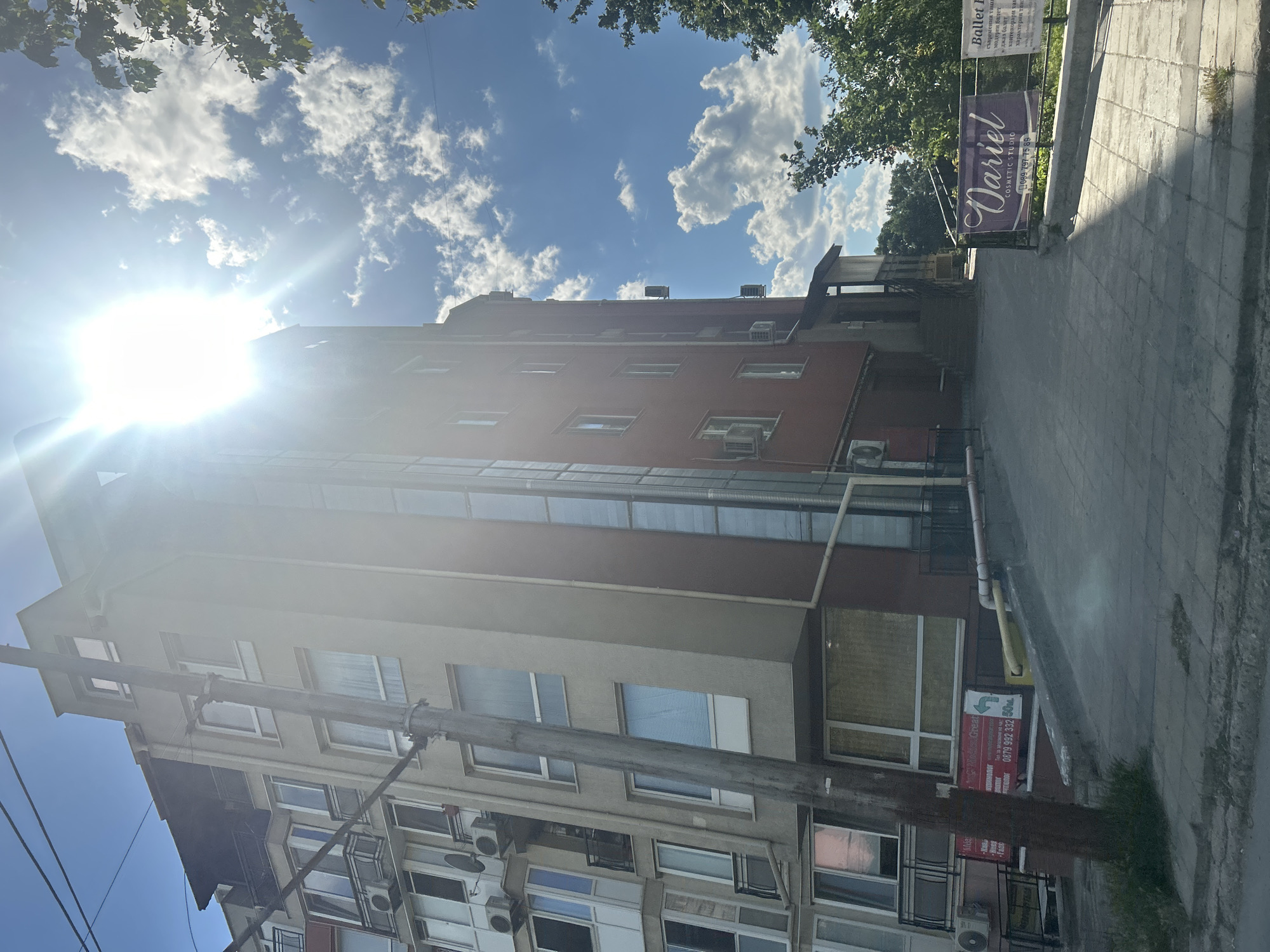 The image shows a multi-story building in Varna, Bulgaria, with a bright sun and scattered clouds in the sky. The building appears to be a mix of residential apartments and commercial spaces. The left side of the building is lighter in color and features multiple balconies and windows, with air conditioning units visible. The right side of the building is darker red and has a glass facade on the upper floors. A utility pole with wires runs vertically in front of the building.
At the ground level, there are signs for "Dariel Cosmetic Studio" and "Ballet F" on banners. A ramp leads up to an entrance on the right side of the building. The area in front of the building is paved with gray tiles, and there is some greenery and trees to the right. The overall scene suggests a street view in a moderately urbanized area. The sun's glare is prominent, making some parts of the image overexposed. There are no people visible in the image.