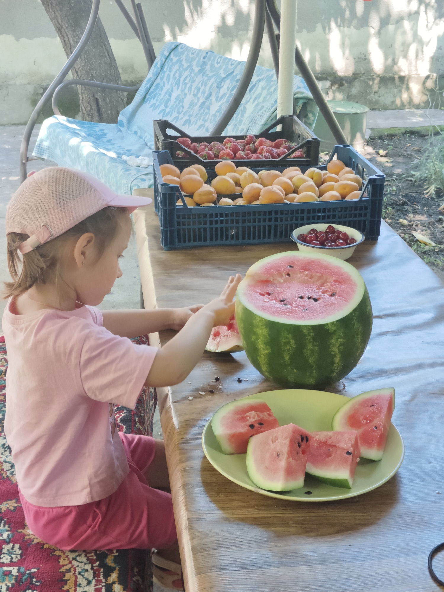 A young girl wearing a pink t-shirt and pink shorts, with a pink baseball cap, sits at a wooden table outdoors, in what appears to be a backyard in Kryvyi Rih, Ukraine. She is reaching towards a partially cut watermelon. Sliced pieces of watermelon are arranged on a light green plate in front of her. Behind her, on the table, are two plastic crates filled with strawberries and apricots, and a small bowl of cherries. The setting includes a swing set with a blue patterned cushion in the background, and a stone wall. The ground is dirt and leaves.