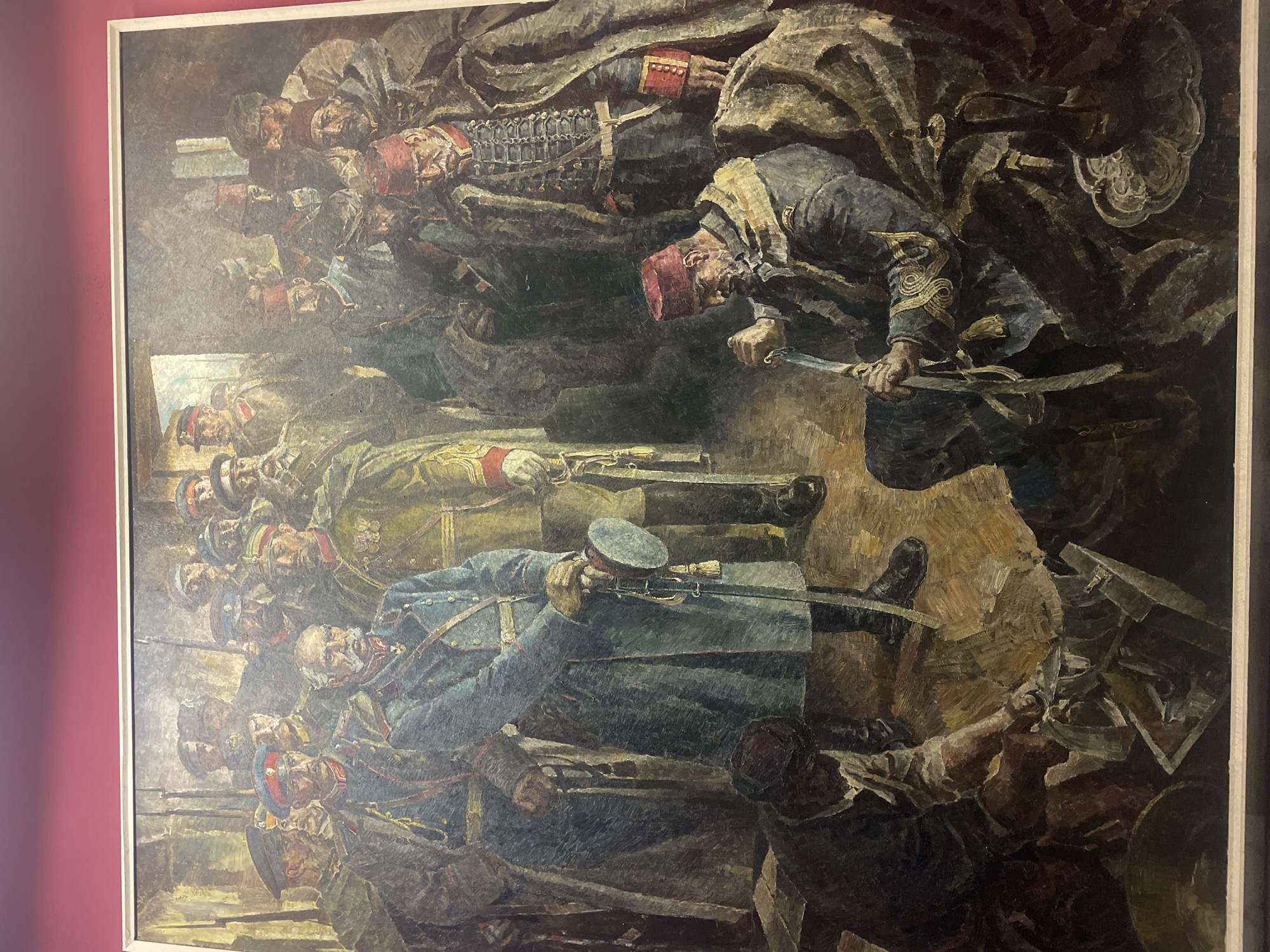 The image is a painting depicting a battle scene. A group of soldiers in blue uniforms and red hats with rifles stand in the lower left. A man in a dark coat and red fez in the upper right wields a sword, positioned above a group of figures clad in darker, more elaborate garb. The painting appears to be located in a museum or gallery, as indicated by the framed artwork and the reddish wall. The painting appears to depict the Siege of Plevna, a key event in the Russo-Turkish War of 1877-1878, which took place in Pleven, Bulgaria.