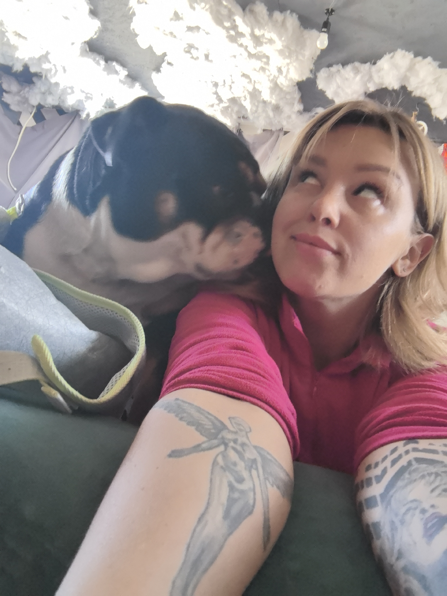 The scene is indoors, located in the city of Noviy, Russia, depicting an adult female and a dog in close physical interaction. The woman has blonde hair and wears a pink long-sleeved shirt. Her left arm displays a grey tattoo of a winged figure, and her right arm shows a dark geometric pattern tattoo. She looks upward and to her right. A black and white dog's head is positioned near the woman's left cheek, indicating nuzzling or licking. A lime-green strap, possibly from a harness or collar, is visible on the dog's shoulder. The ceiling features white, cloud-like decor and a single bare light bulb in a dark fixture. The immediate foreground is a dark, upholstered surface.