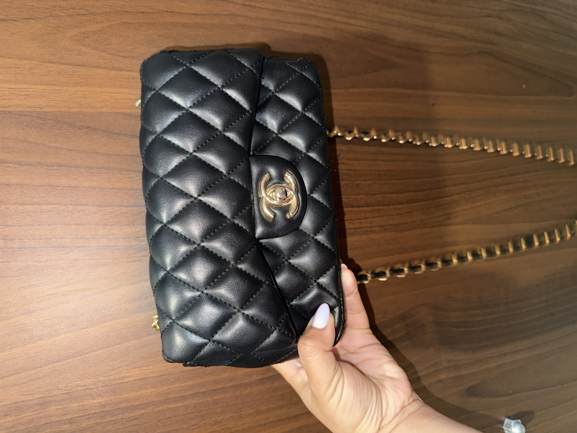 A black, quilted Chanel handbag with a gold chain strap is held in a person's hand against a wooden surface. The bag's CC logo is visible. The person has a light-colored nail polish on their thumb. Part of the person's arm and what appears to be a metal bolt are visible in the lower right corner. The location is Unknown, Unknown.
