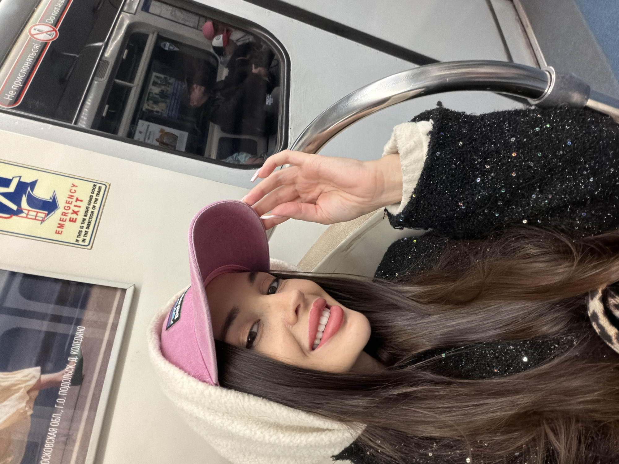 The photograph shows a woman wearing a pink baseball cap and a black jacket with white trim. She is smiling and looking directly at the camera, with her hand raised near her face. The photograph was taken inside a train, as evidenced by the window and handrail visible in the background, and the signs in Russian indicating "Emergency Exit" and "Do not lean." A poster on the wall references "Moscow Oblast, Podolsk, D. Koledino," suggesting this is a metro or train line in or near Moscow, Russia.