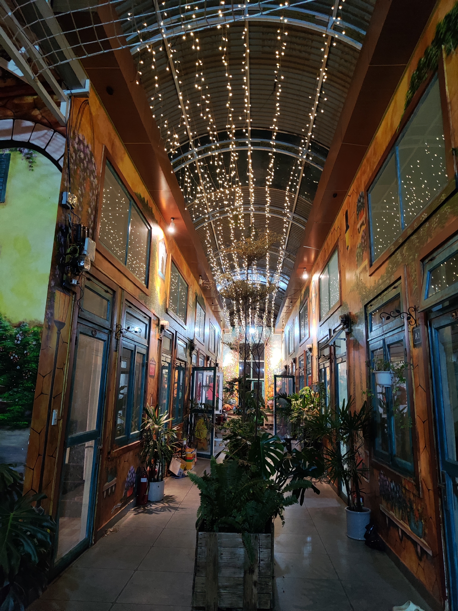 The image depicts an indoor alleyway in Da Lat, Vietnam, illuminated by strings of lights suspended from the arched ceiling. The walls on either side feature faux facades of buildings with doors and windows, painted with murals. Plants in pots and planters line the passageway. There are no people immediately visible. The floor is tiled.