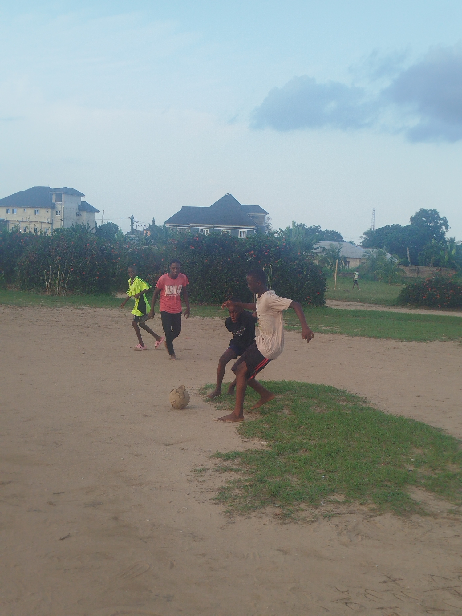 The scene depicts a group of four young boys playing soccer on a sandy field in Ikot Mbonde, Nigeria. Three boys are clustered around the soccer ball. One boy in a yellow shirt and another in a pink shirt run in the background. A house, buildings, and green foliage are in the background.