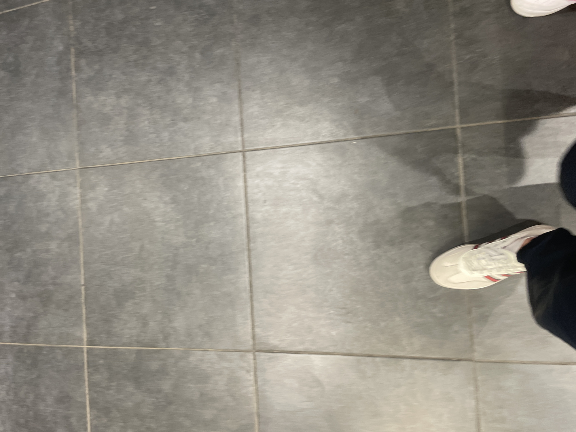 The scene is set in Ihtiman, Bulgaria, and features a ground-level, high-angle perspective of a tiled floor. The floor is constructed from large, rectangular dark grey tiles with subtle variations in shading, exhibiting a matte finish and some signs of wear. Light-colored grout lines form a distinct grid pattern across the entire visible surface.
In the lower portion of the image, the feet of two individuals are present. One foot, positioned towards the lower left, is clad in a dark pant leg and a cream-colored sneaker with prominent red and white stripes running diagonally across its side. The tip of a second, light-colored shoe is partially visible in the extreme lower right corner of the frame. Two elongated shadows are cast on the tiled floor, originating from the lower edge and extending upwards, indicating an overhead or back-lighting source. The arrangement of the feet suggests a momentary pause or motion within an indoor environment.
