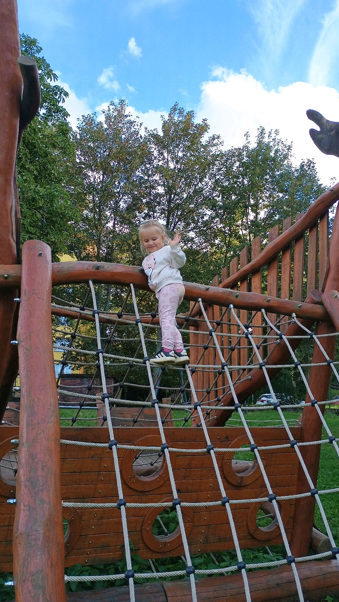 A young girl is standing on a rope net on a wooden playground structure that resembles a ship. She is smiling and waving her right hand. The girl is wearing a light-colored sweatshirt and patterned leggings. The playground is surrounded by trees and a grassy area. The sky is blue with some clouds. The playground is located in Sokolov, Czechia.