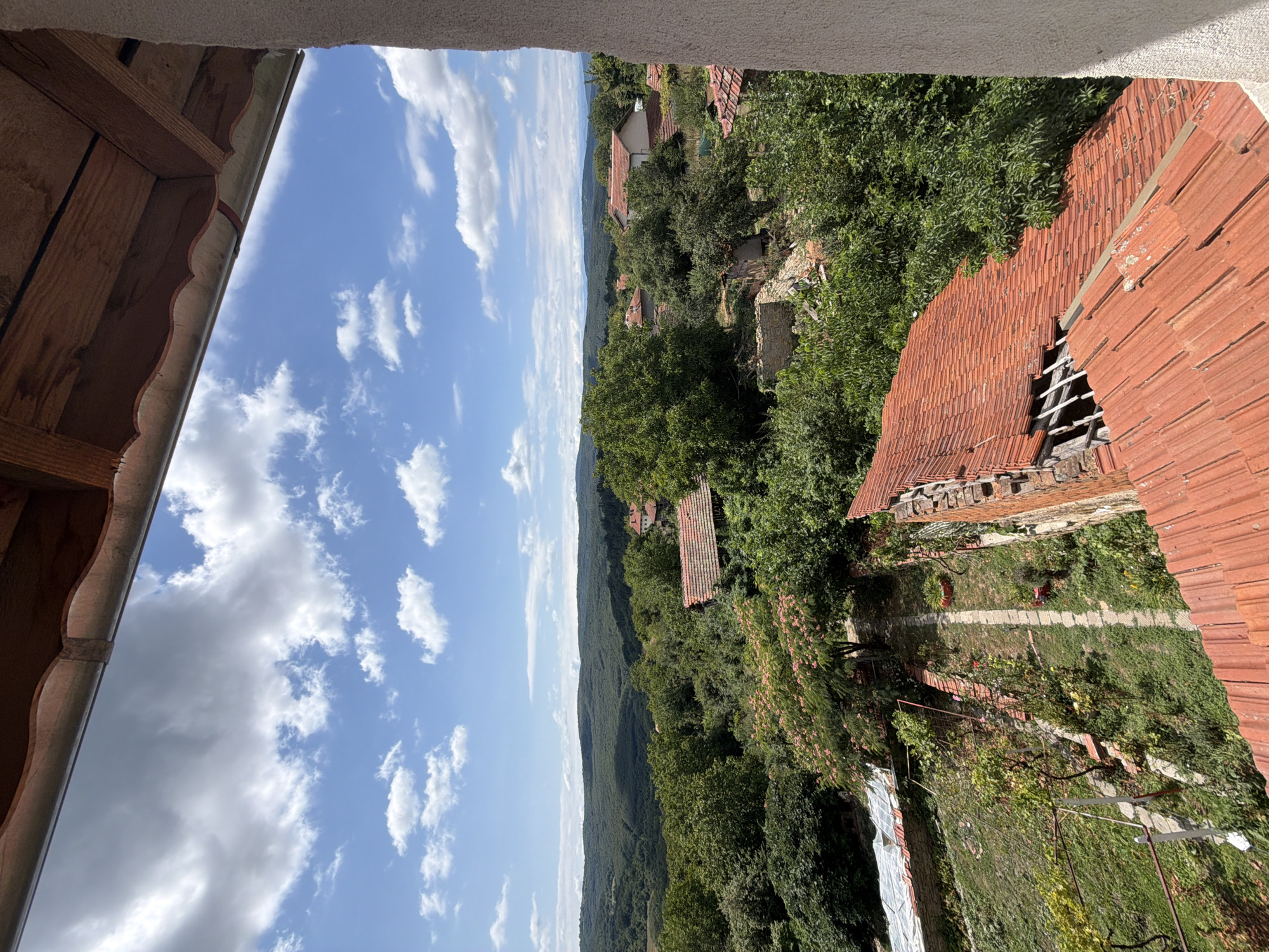 This is a daytime, high-angle photograph taken from a structure looking out over a rural landscape in the vicinity of Gorno Pavlikene, Bulgaria. The foreground is dominated by the roofline of a building with red terracotta tiles, some of which appear damaged. Beyond this, the scene unfolds into a village characterized by similar red-tiled roofs and lush green vegetation. Trees are abundant, with a prominent flowering tree with pink blossoms in the mid-ground. Hills covered in dense forest form the distant backdrop. The sky is blue with scattered white cumulus clouds. A pathway with stone slabs is visible in the lower left quadrant, leading into a garden area with some white plastic sheeting covering what appears to be a greenhouse or protective covering. There are no people or animals visible in the image. The overall impression is one of a sunlit, tranquil rural setting.