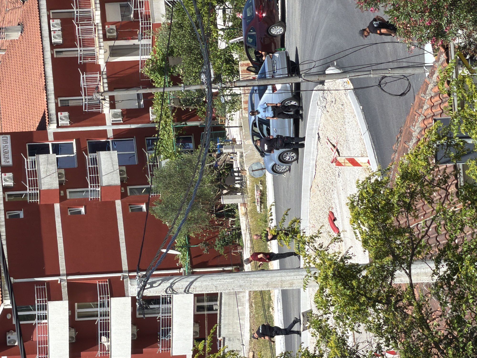 The image depicts a bright, sunny daytime street scene, likely in Kubasi, Montenegro, featuring a notable police presence. In the center-right, two police officers stand near a silver sedan with its front driver's side door open. One officer wears a light blue shirt and dark pants, while the other is in a darker uniform. A third individual, also in dark attire, is further back near the silver car. Behind it, a dark red sedan is parked.
On the left side of the frame, a prominent multi-story building with reddish-brown walls and white trim features numerous balconies with white railings, windows, and external air conditioning units. The text "APARTMENTS GOLD" is clearly visible on its side. The building is topped with a terracotta-tiled roof.
Several other individuals are present on the pavement and street. One person in a red shirt and dark pants stands on the pavement, while another in a dark t-shirt and cap is further down the road. A utility pole with numerous cables runs vertically through the scene. A red and white striped barrier lies on the ground near the silver car. Green trees and bushes, some with pink flowers, are scattered throughout, and strong sunlight casts clear shadows, indicating good weather. The scene suggests an official incident or police activity on a residential or commercial street.