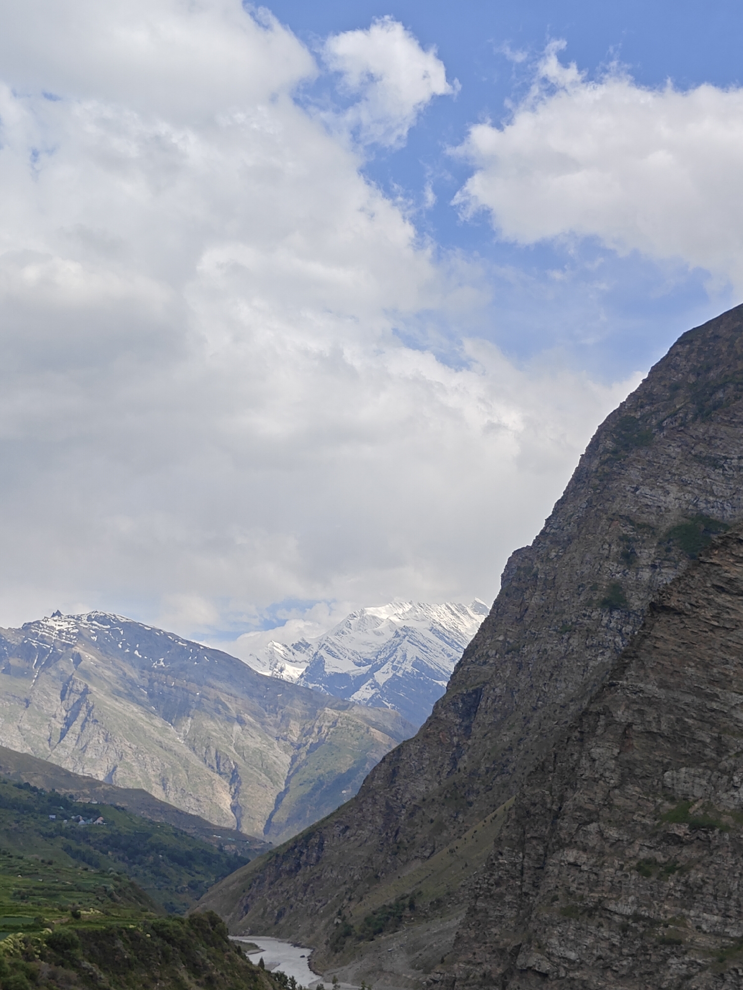 The image depicts a mountainous landscape with a river flowing through a valley. The foreground is dominated by a steep, rocky mountain face on the right. In the mid-ground, a wide river cuts through the valley, flanked by green, terraced hillsides. In the background, snow-capped mountains rise majestically under a partly cloudy sky with patches of blue. The scene is devoid of any people or human activity, focusing solely on the natural environment. The location is identified as Kirting, India, a region known for its dramatic mountain scenery.