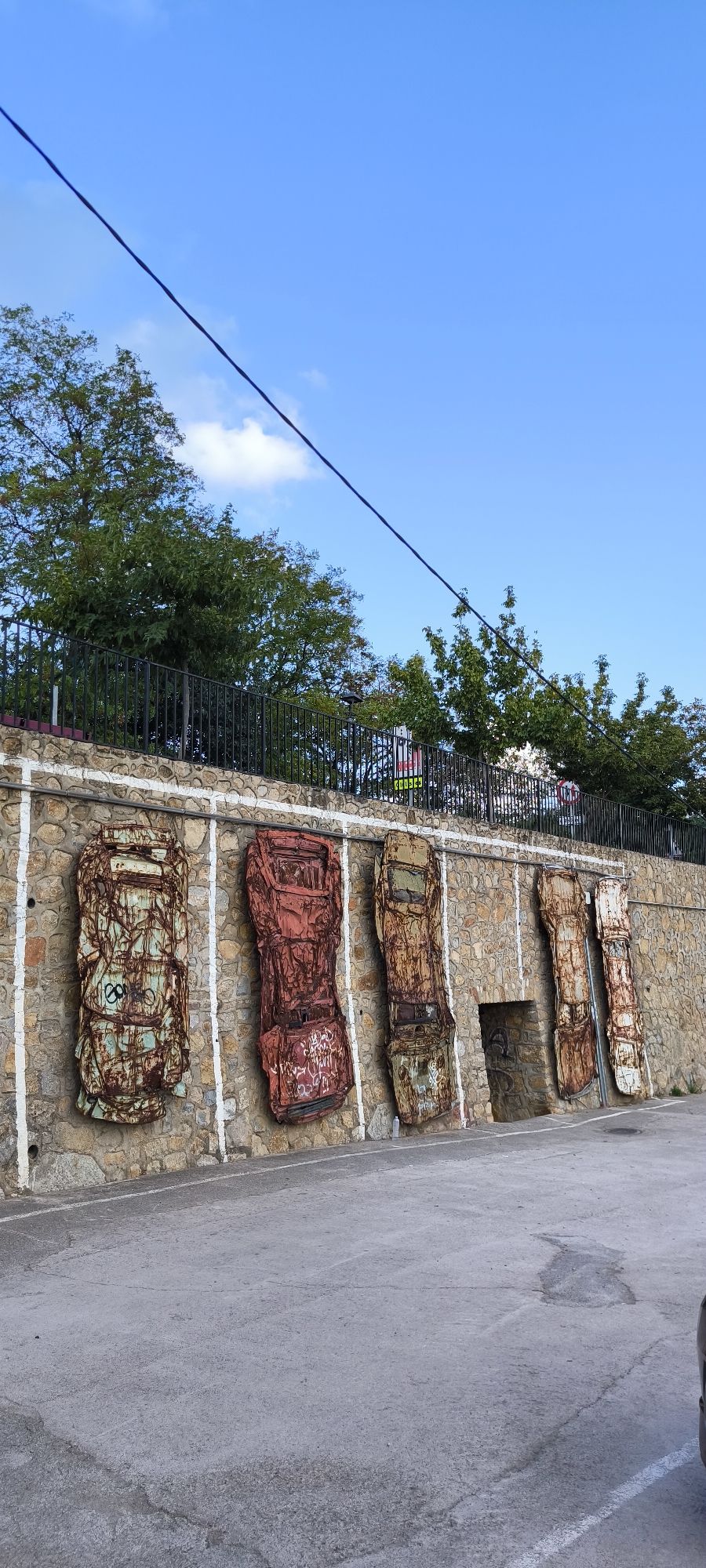 In the city of Fanzara, Spain, a stone wall displays a row of five flattened, rusted vehicles mounted vertically. The vehicles, appearing to be old car bodies, are painted in various states of decay, with the leftmost one exhibiting a turquoise hue and graffiti. White lines painted on the wall suggest parking spaces. Behind the wall, a black metal fence encloses trees, and a blue sky with scattered clouds forms the background. A black wire stretches diagonally across the upper portion of the image. To the right of the vehicles, a small, dark arched opening is visible in the wall. The foreground consists of a paved surface, likely a parking lot.