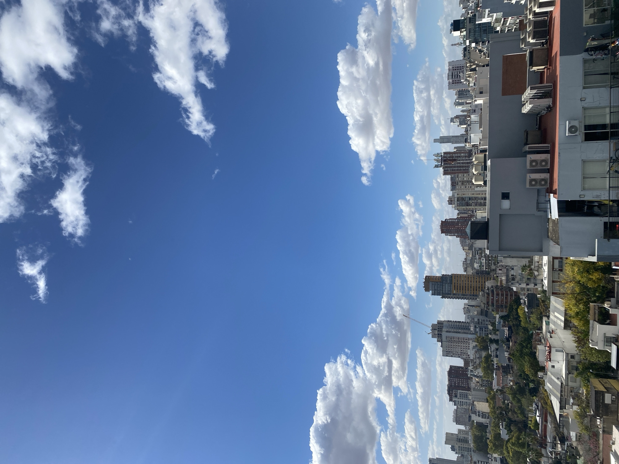 A cityscape in Buenos Aires, Argentina, is captured under a blue sky dotted with fluffy white clouds. The architecture varies, with a mix of high-rise buildings and smaller residential structures interspersed with trees. Air conditioning units are visible on the sides of some buildings. The scene appears to be taken from an elevated position, possibly from another building.