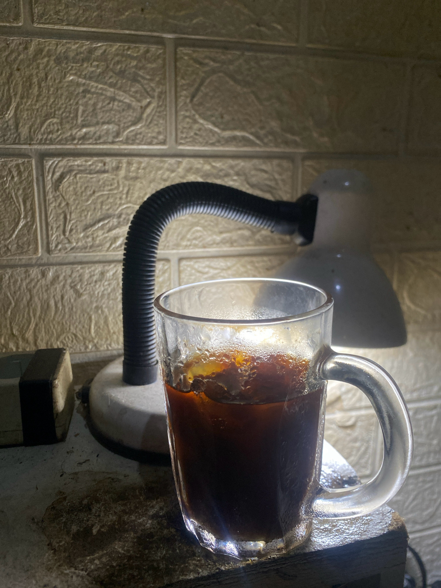 A glass mug filled with iced coffee sits on a textured surface in front of a white brick-patterned wall. A desk lamp with a flexible black hose is positioned behind the mug, its white shade illuminated by the light. To the left of the mug, a small, dark rectangular object with a white stripe is visible. The overall lighting suggests a dimly lit indoor environment, possibly a home or office, in Batam, Indonesia, given the prompt's context. There are no people or animals present in the image. The focus is on the still life arrangement of the coffee and desk lamp.