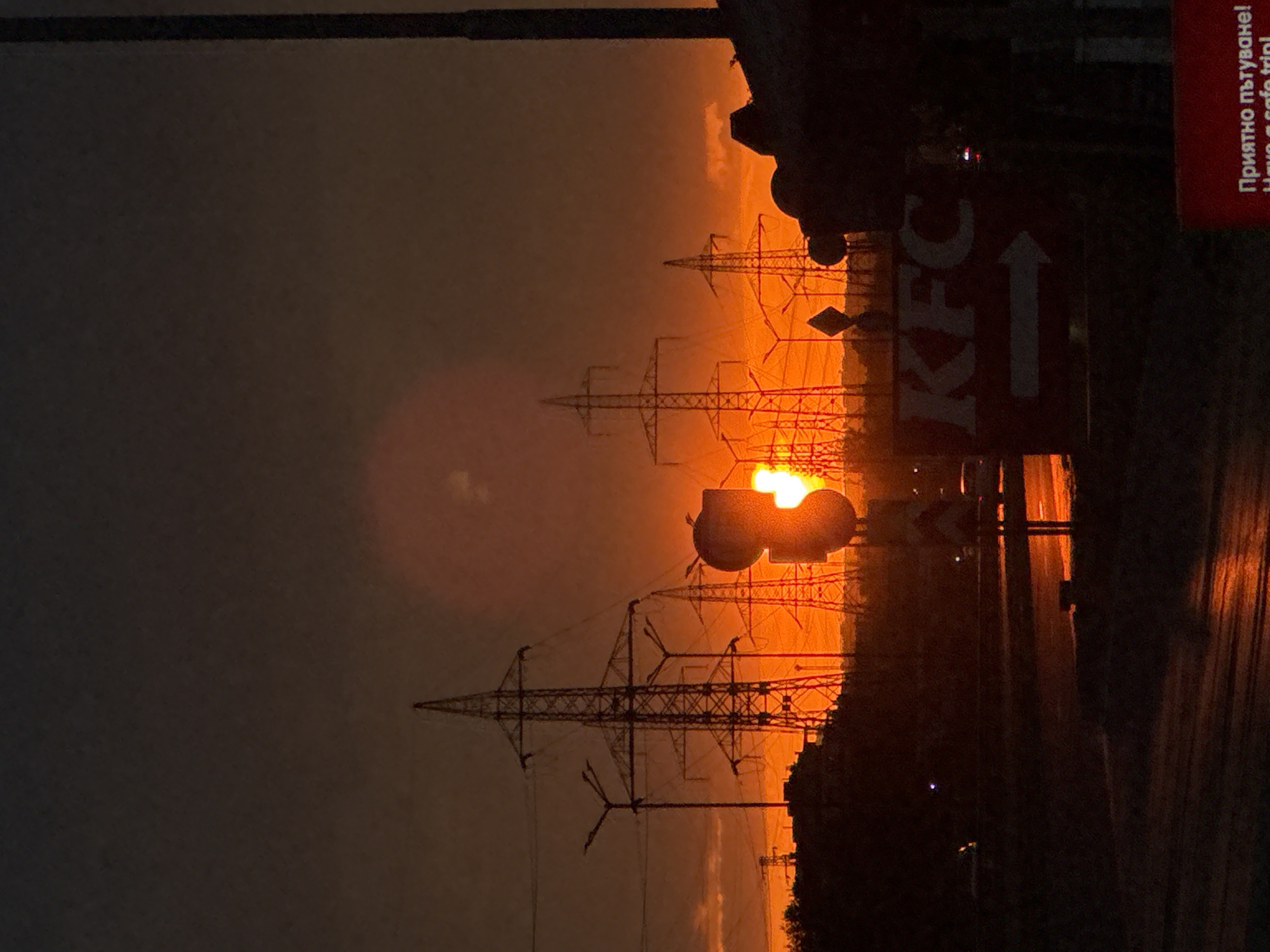 The image shows a sunset in Sofia, Bulgaria. In the foreground is a KFC sign with an arrow pointing towards the restaurant. In the background, there are power line towers silhouetted against the orange sky. A sign with Bulgarian text is present on the right edge of the image. The scene is viewed from a ground-level perspective, with a road visible in the lower right corner, reflecting the orange light of the sunset.