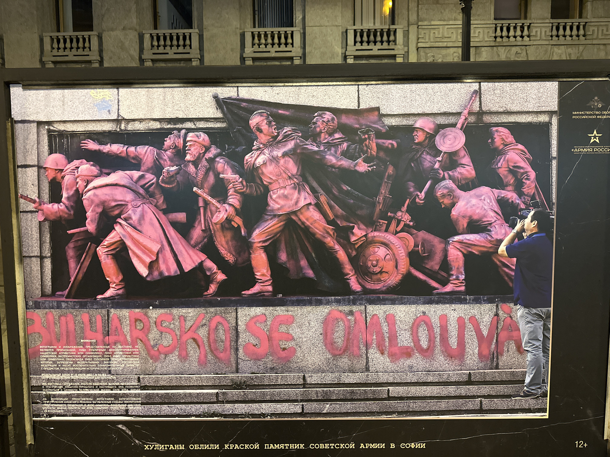 The image shows a large, rectangular sign depicting a Soviet-era monument. The monument features a relief of soldiers in action, cast in what appears to be bronze or a similar material. The relief is depicted with pink paint splashed over it, and graffiti in what seems to be Bulgarian is painted in large pink letters at the bottom of the monument's base. To the right of the sign stands a man in a blue shirt and light-colored pants, facing towards the sign and holding what appears to be a smartphone up to his face, likely taking a photograph. The sign is part of an outdoor display, with a black frame and informational text in Russian at the bottom and to the right, and logos of the Russian Ministry of Defense, and "Army of Russia". The sign bears the text "ХУЛИГАНЫ ОБЛИЛИ КРАСКОЙ ПАМЯТНИК СОВЕТСКОЙ АРМИИ В СОФИИ" ("Hooligans poured red paint on the Soviet Army monument in Sofia"). The location is not St. Petersburg, Russia, but is presumably Sofia, Bulgaria. The background shows a building facade with architectural details, including balconies. The lighting is ambient and suggests an outdoor setting at dusk or in the evening.