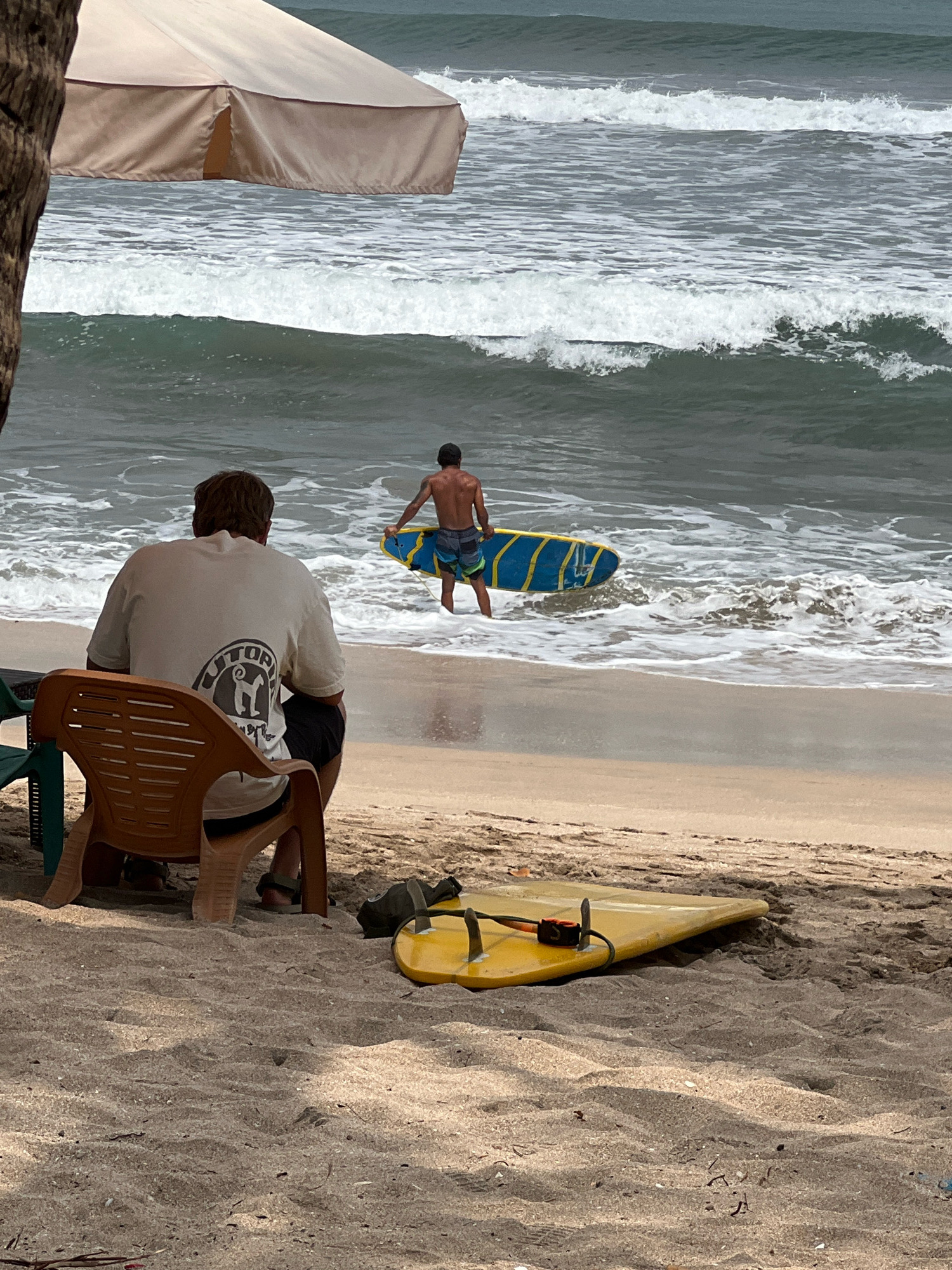 In Kuta, Bali, Indonesia, a beach scene unfolds under a partially visible tan umbrella. In the foreground, a person sits in a tan plastic chair, facing away from the camera and towards the ocean. They are wearing a light-colored t-shirt with a circular logo on the back and dark shorts. A yellow surfboard rests on the sand in front of the chair.
Further out, closer to the water's edge, another person, shirtless and wearing patterned swim trunks, walks into the ocean carrying a blue and yellow surfboard. Waves are breaking on the shore, creating white foam. The sand appears damp near the water and dry further up the beach. The ocean water is a dark, greenish-gray.